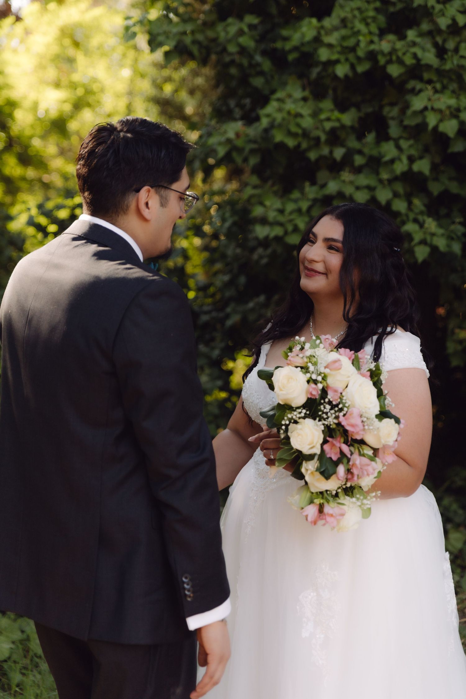 Smiling bride in white wedding dress holding pink roses bouquet faces groom in black suit among green trees.