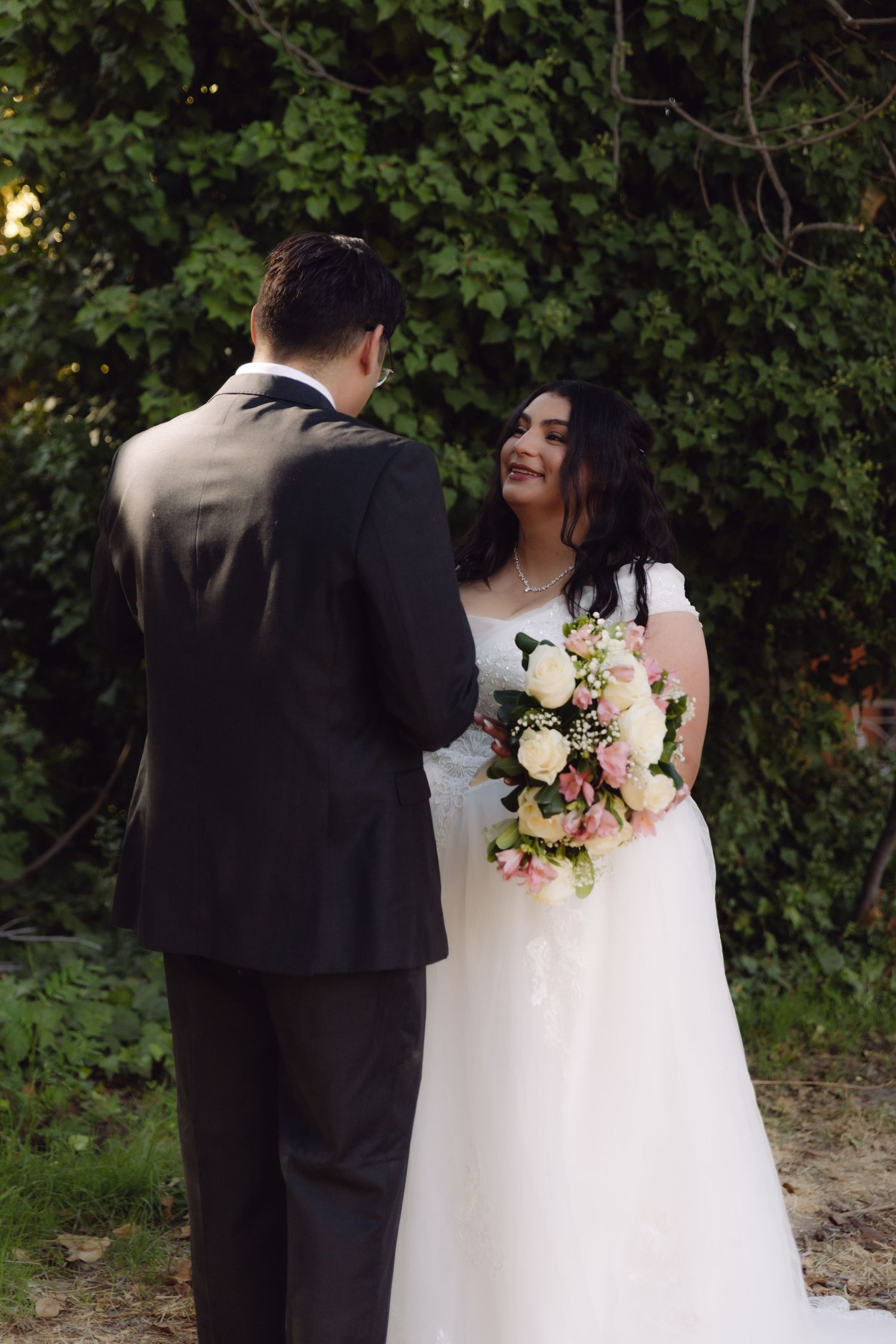 Bride smiling at groom in lush garden setting, holding elegant pink and white floral bouquet in white gown.