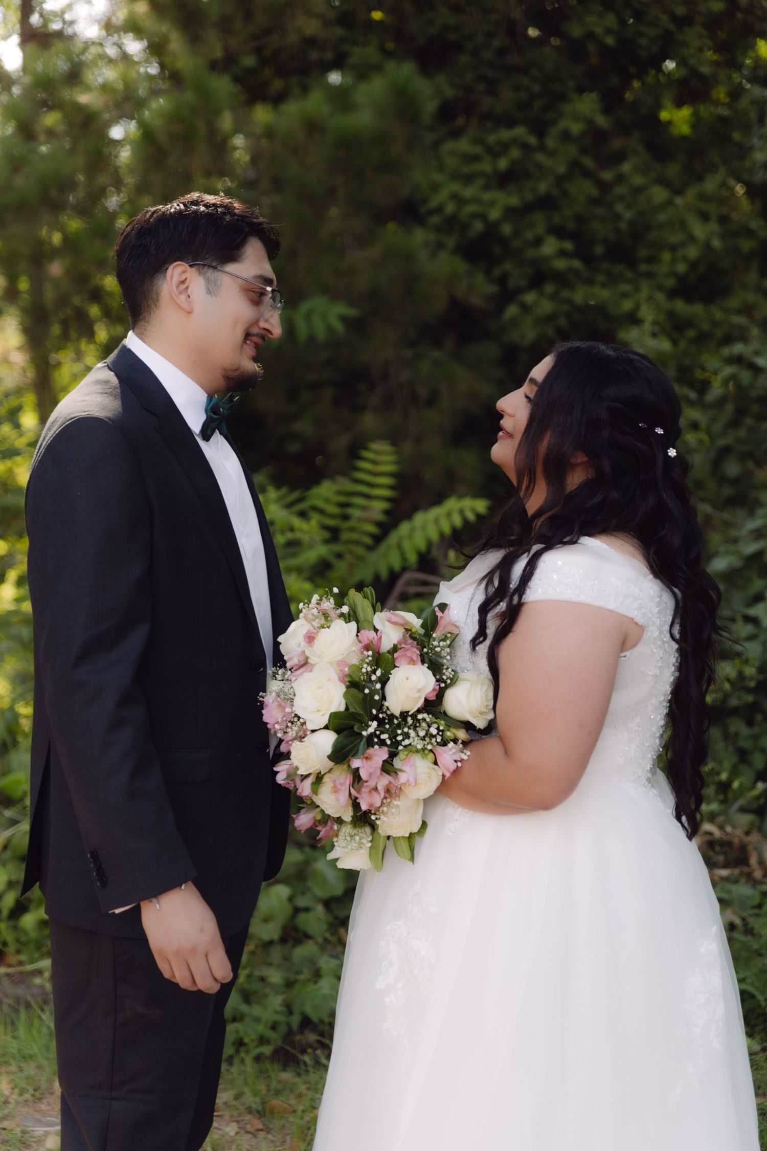 Bride and groom smiling at each other in garden, bride holding pink roses, groom wearing black bow tie.