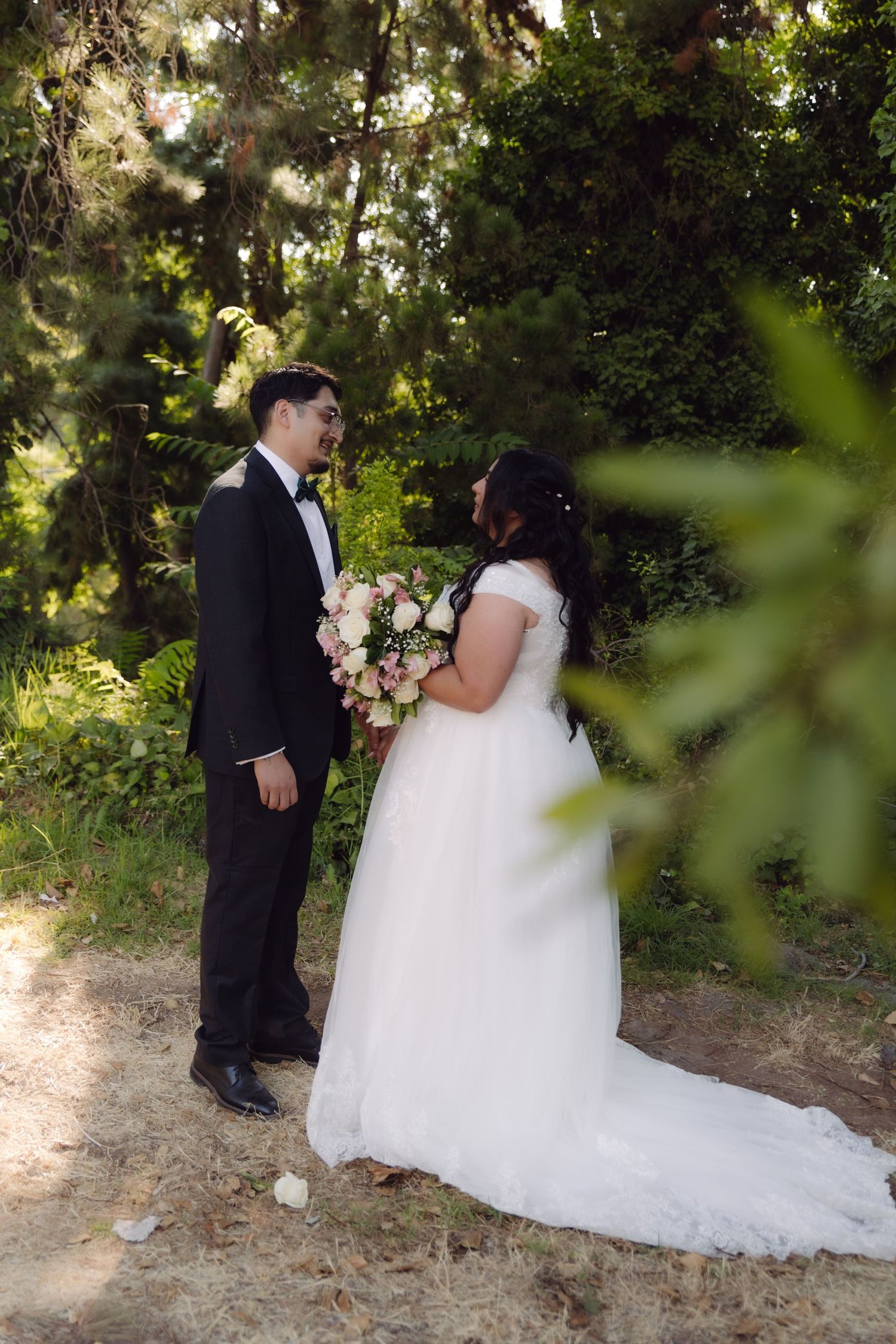 Wedding couple standing face to face in sunlit forest, bride holding bouquet in white dress with long train.