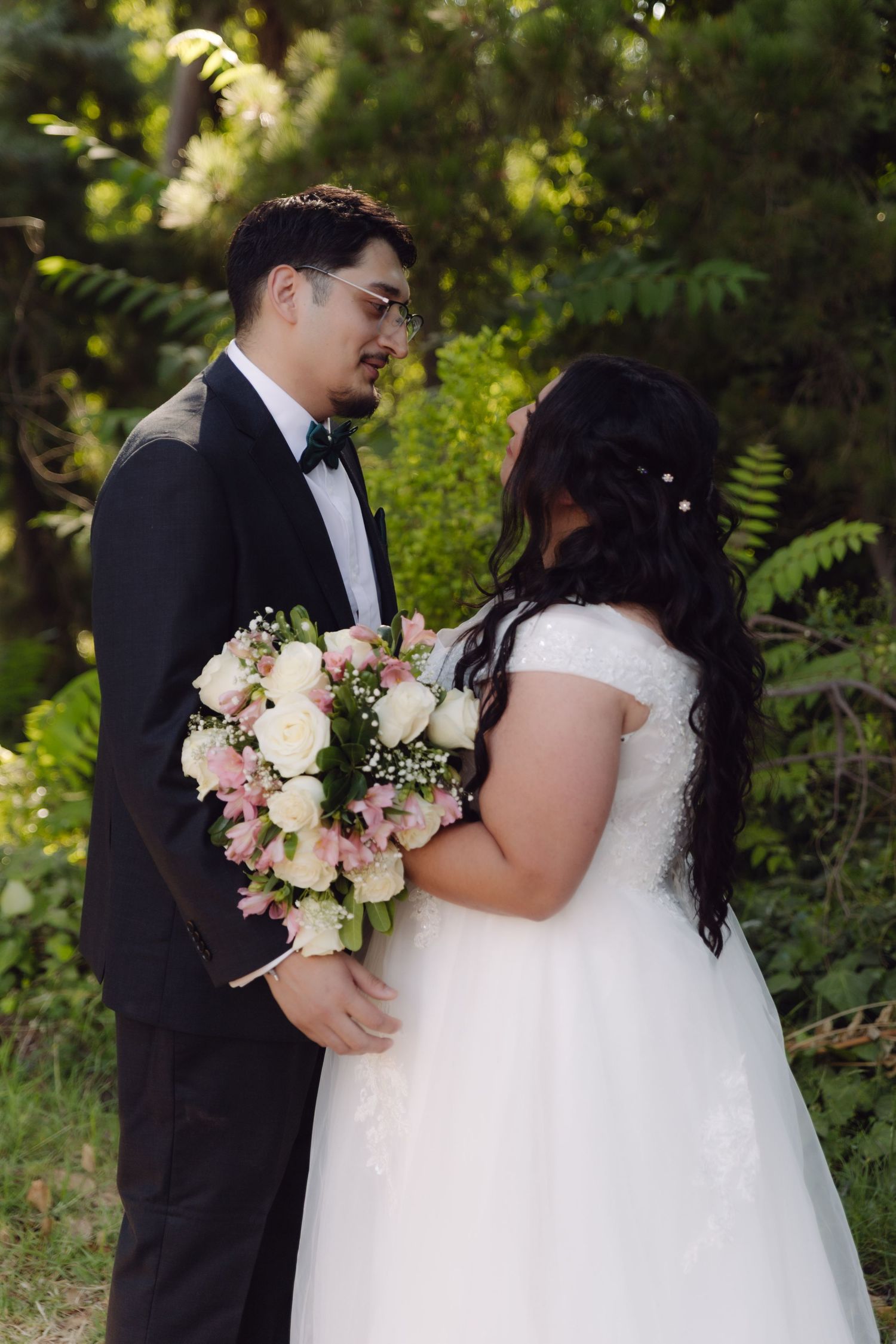 Groom in black tuxedo and bride in white lace gown share intimate moment holding pink and white bouquet.