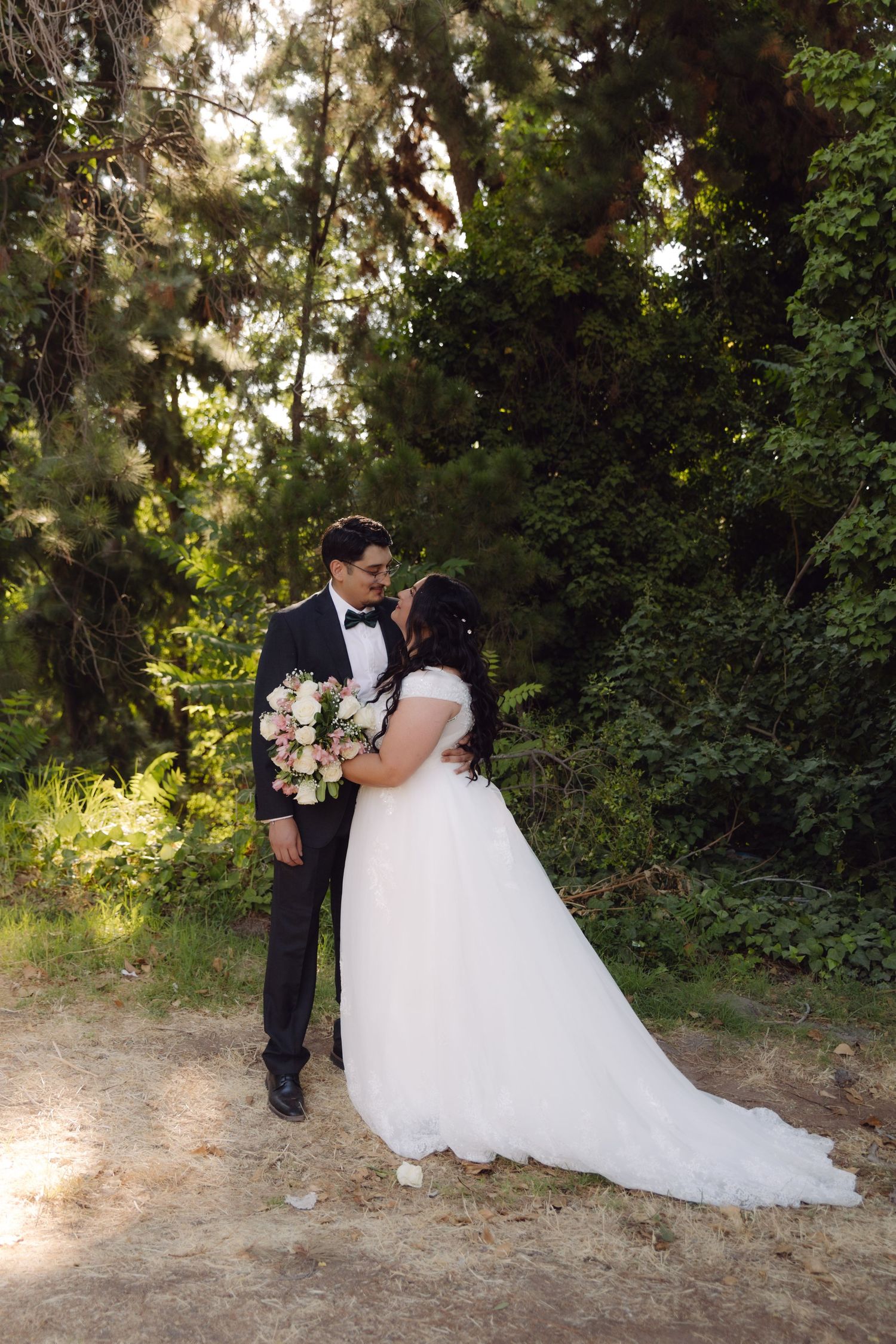 Newlyweds sharing a kiss in a sunlit forest path, bride in white gown holding pink and white bouquet.