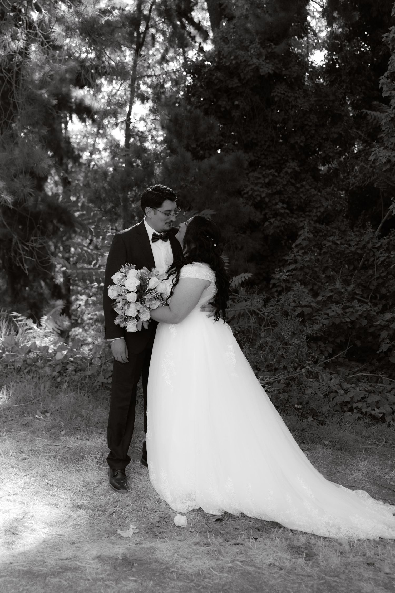 Black and white photo of newlyweds kissing in forest, bride in flowing white gown holding floral bouquet.