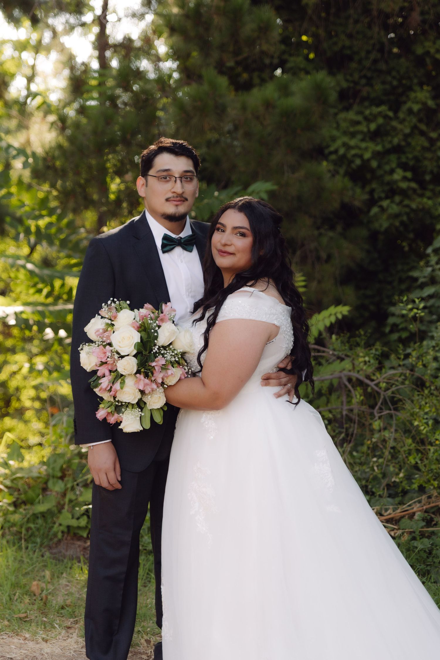 Bride and groom posing together in garden, groom in black tuxedo, bride holding pink and white floral bouquet.