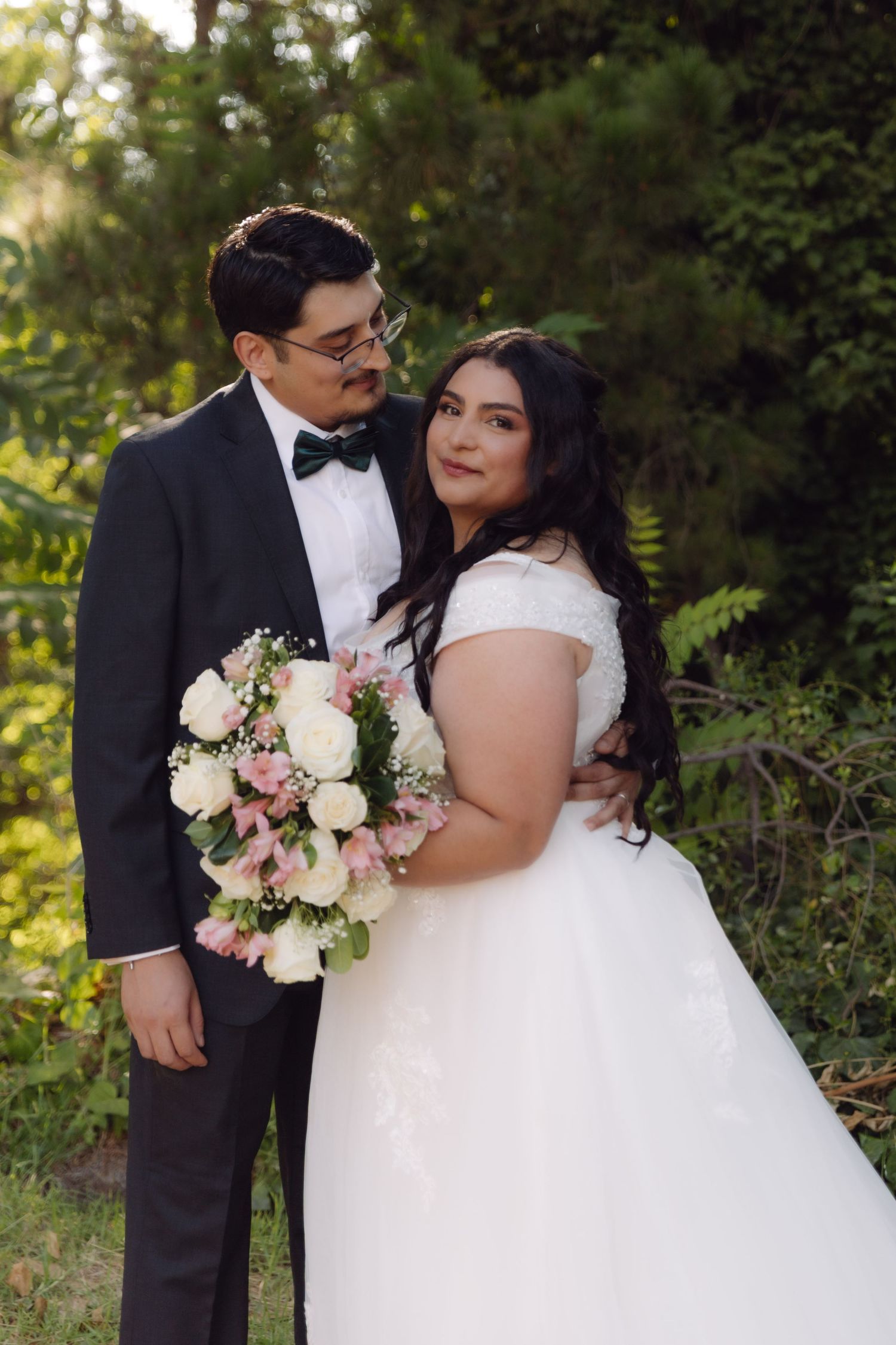 Groom in black tuxedo embracing bride in white off-shoulder gown holding pink bouquet in lush green setting.