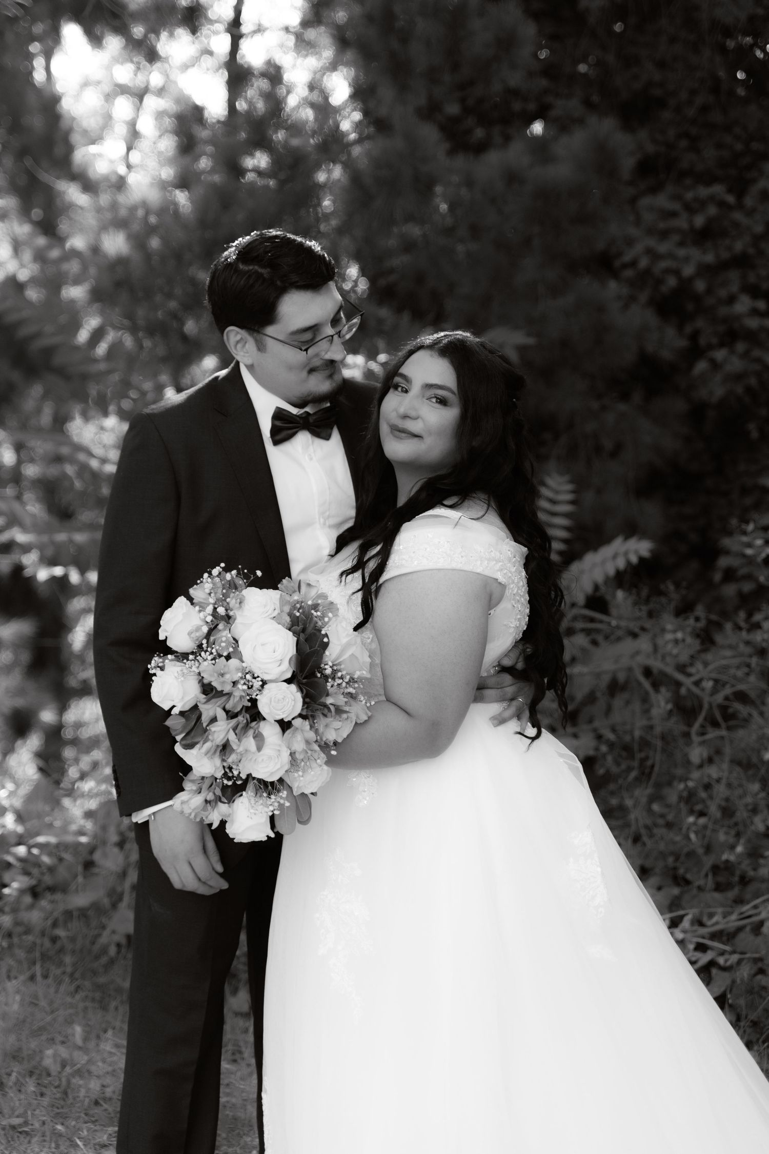Black and white wedding portrait of bride and groom embracing in lush garden setting.