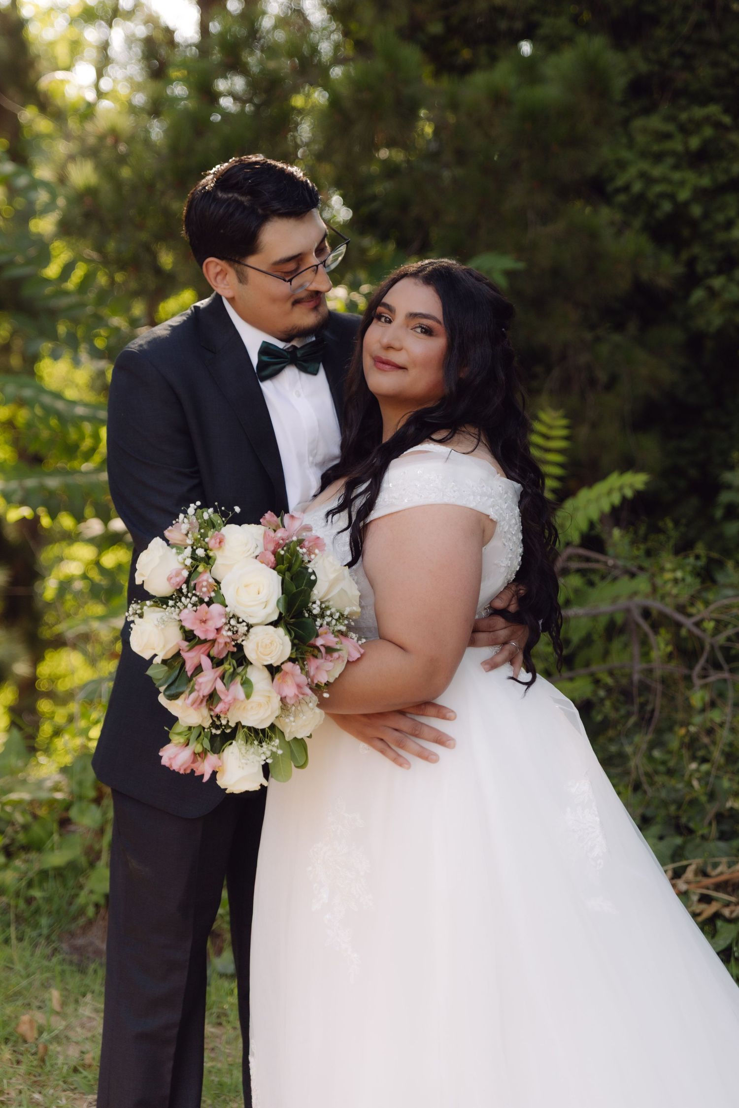 Bride and groom sharing romantic embrace outdoors holding pink and white wedding bouquet.