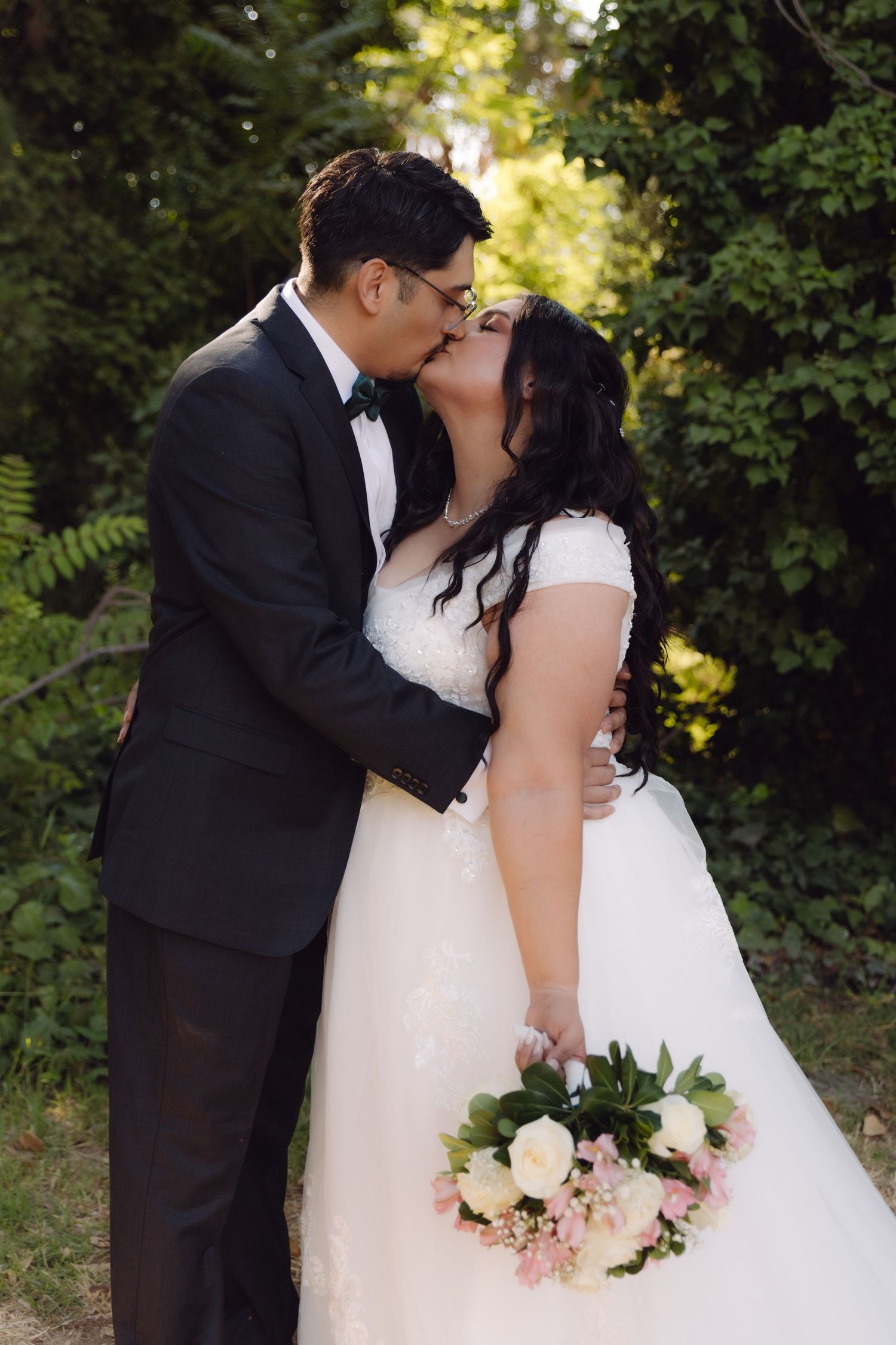 Bride and groom sharing a kiss outdoors surrounded by green foliage on wedding day.