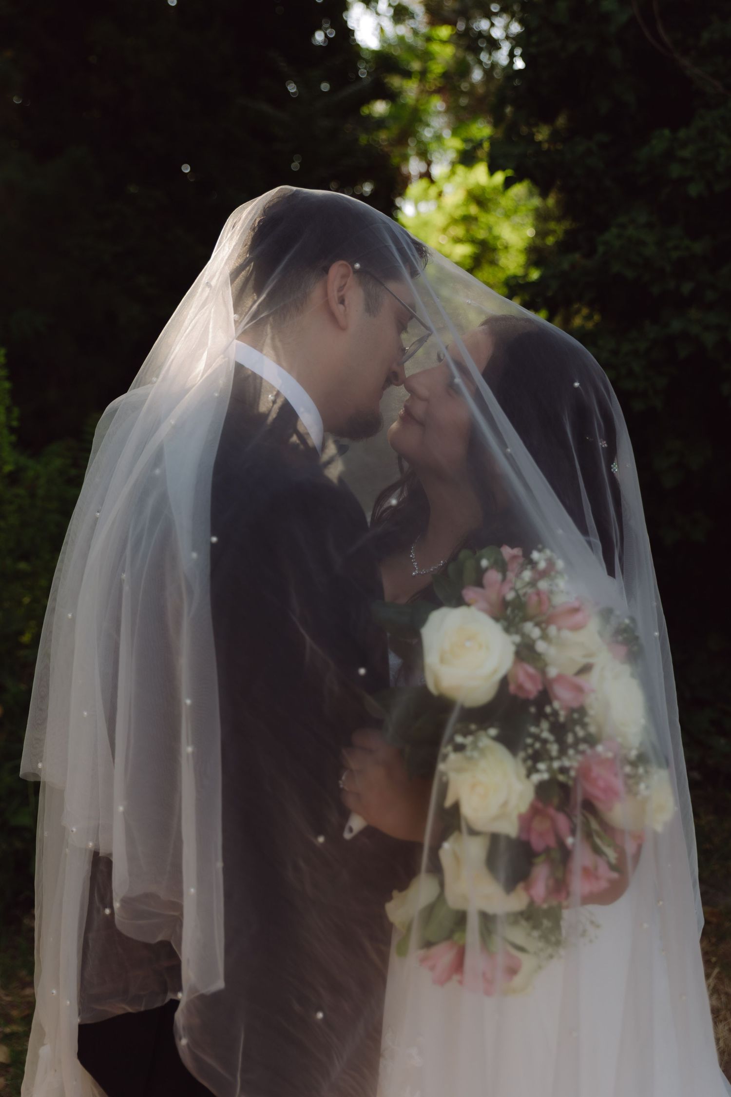 Romantic wedding couple kissing under bridal veil while holding pink and white bouquet.