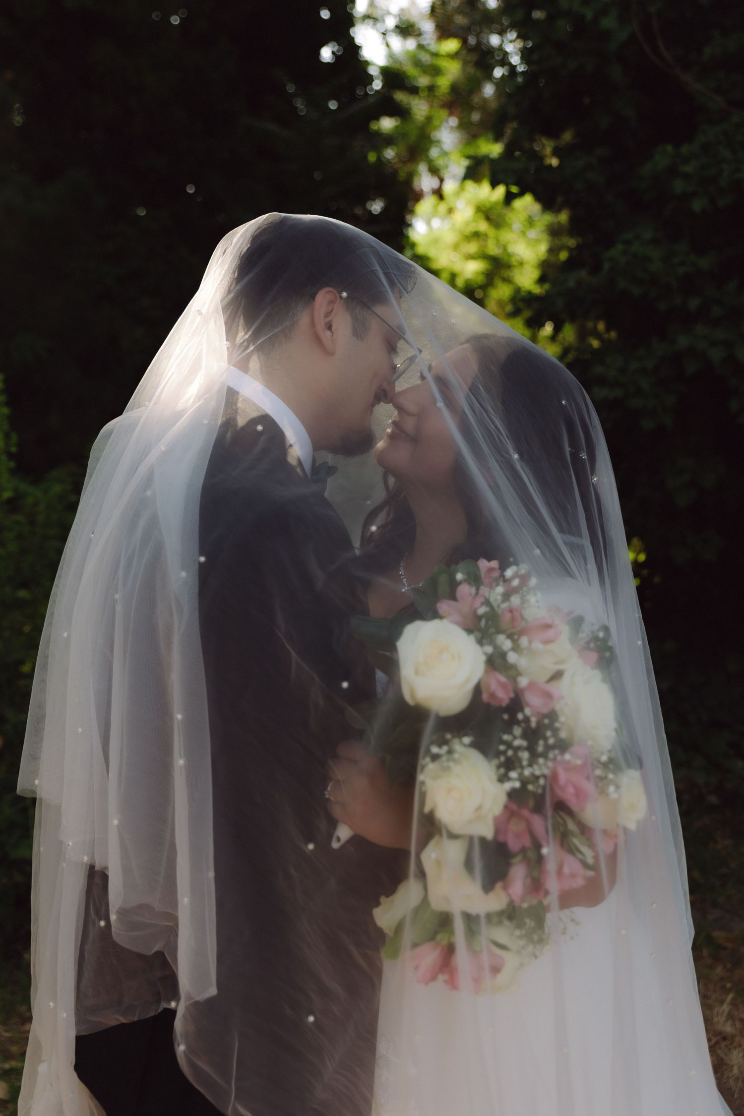 Wedding couple nearly kissing under a sheer veil outdoors, bride holding a pink and white floral bouquet.