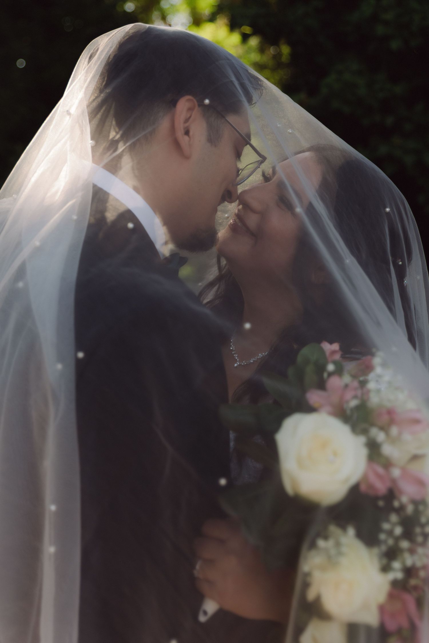 Bride and groom sharing intimate moment under wedding veil in sunlit garden setting.