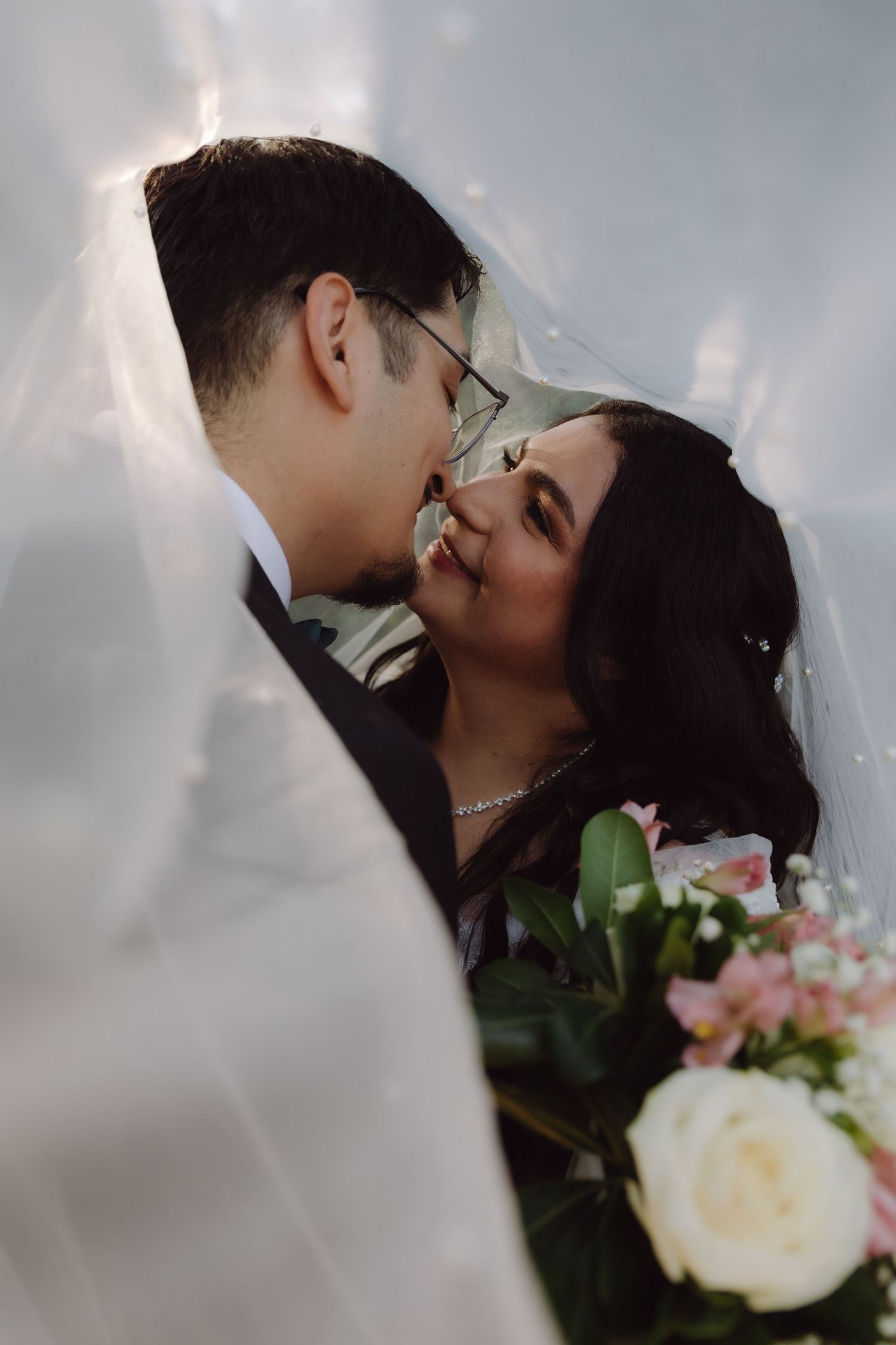 Close-up of bride and groom sharing a kiss under wedding veil with bouquet visible.