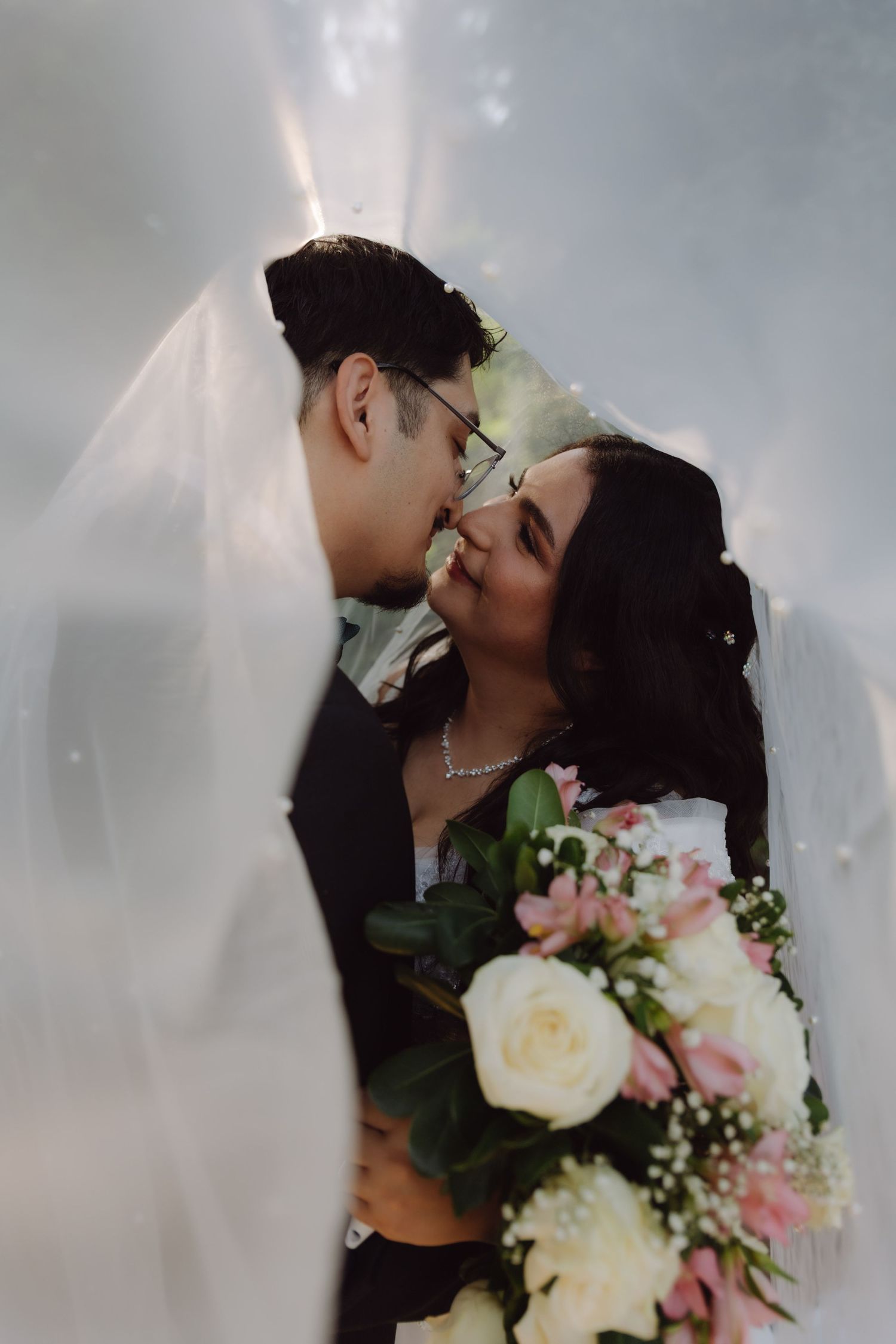 Bride and groom share a romantic kiss under a flowing wedding veil, bride holding a pink and white bouquet.