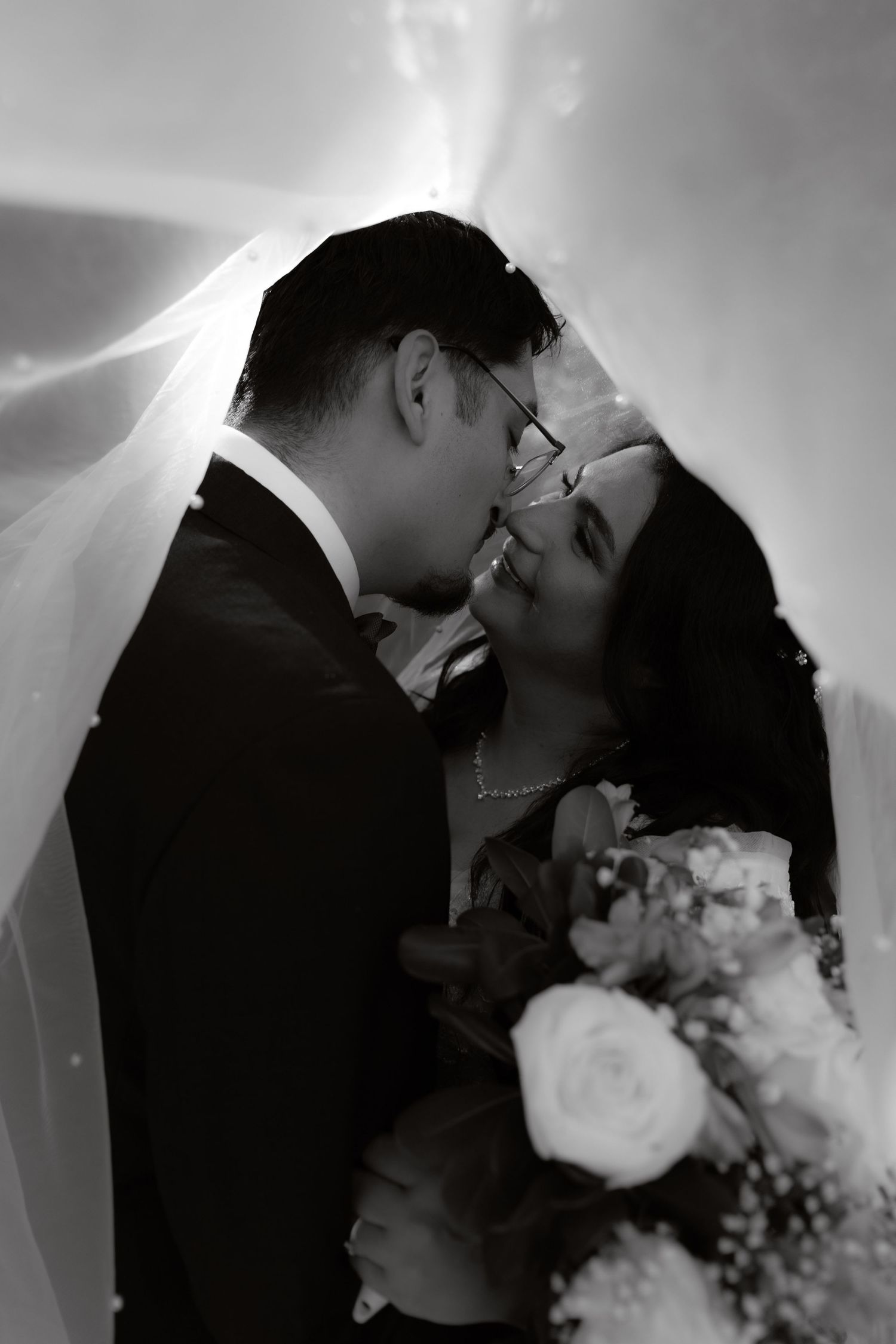 Black and white photo of bride and groom kissing under a wedding veil, bride holding a bouquet of roses.
