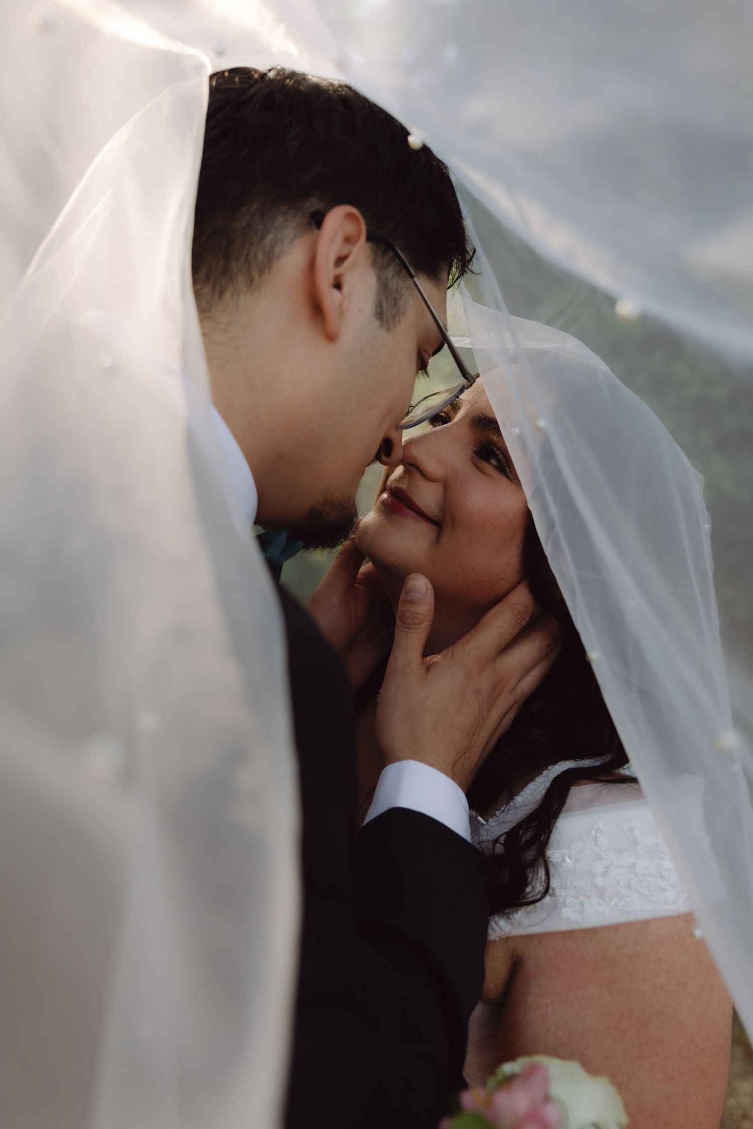 Groom tenderly kissing bride under flowing wedding veil in close romantic portrait.