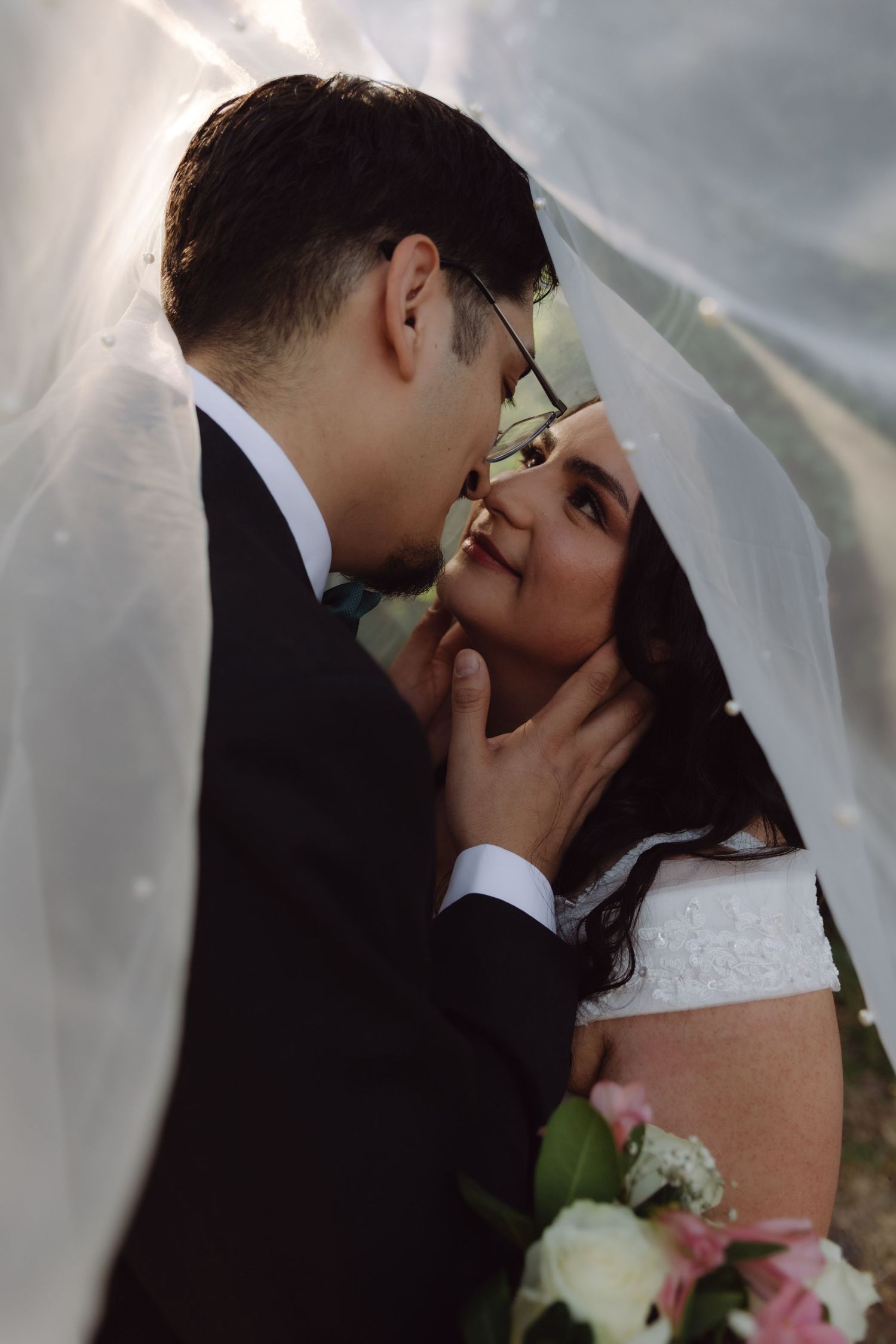 Groom tenderly holds bride's face under a flowing wedding veil as she gazes up at him lovingly.