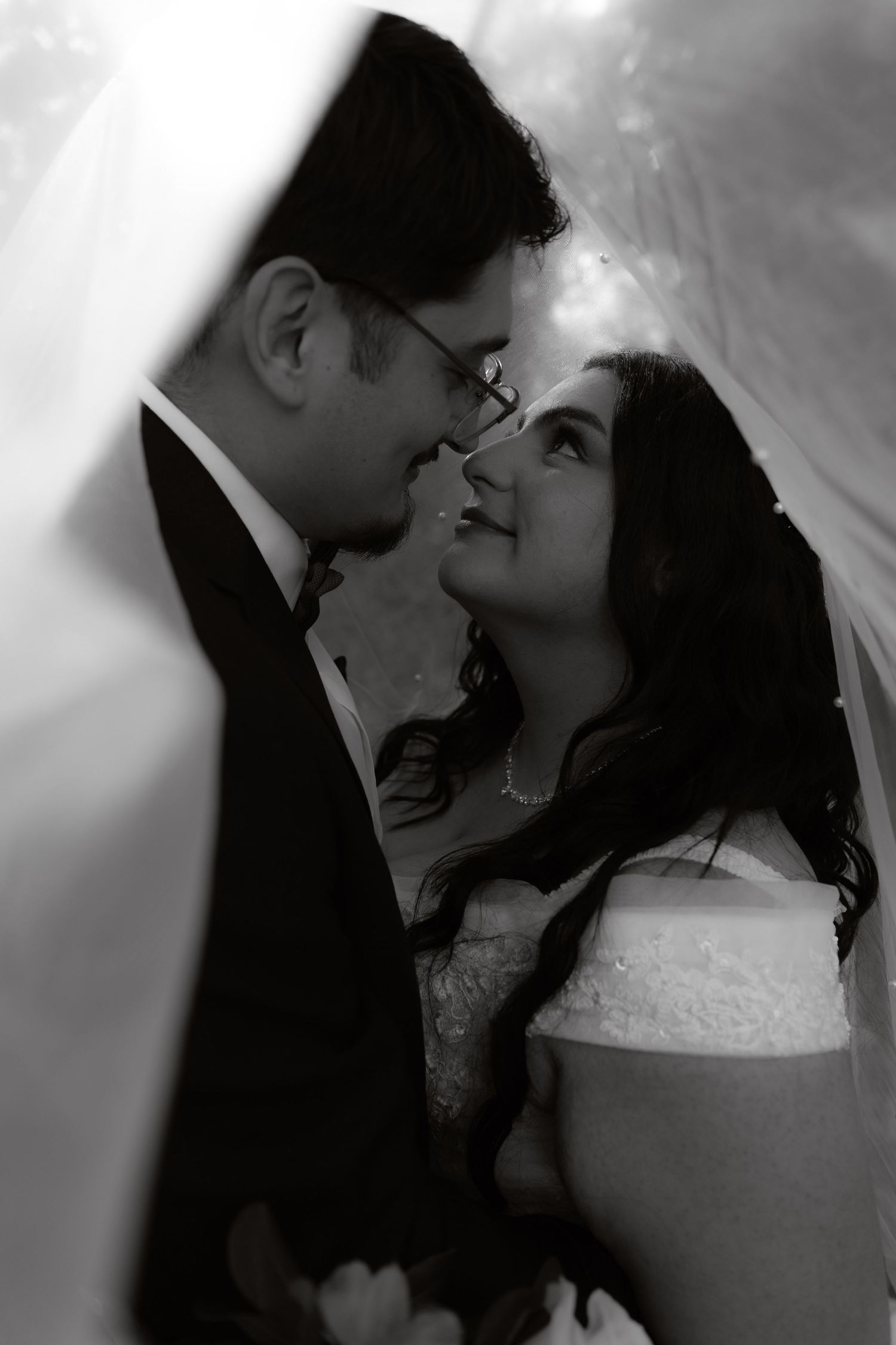 Black and white romantic portrait of bride and groom gazing at each other under a flowing wedding veil.