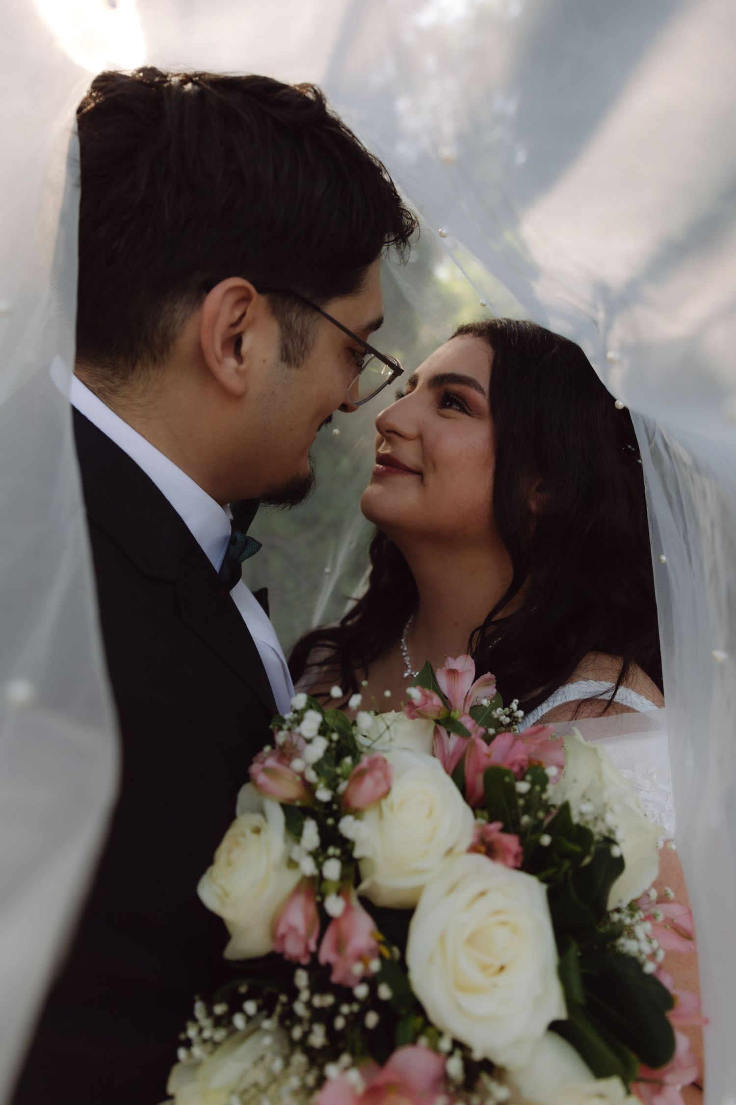 Bride and groom share an intimate moment under a wedding veil, bride holding pink and white roses.
