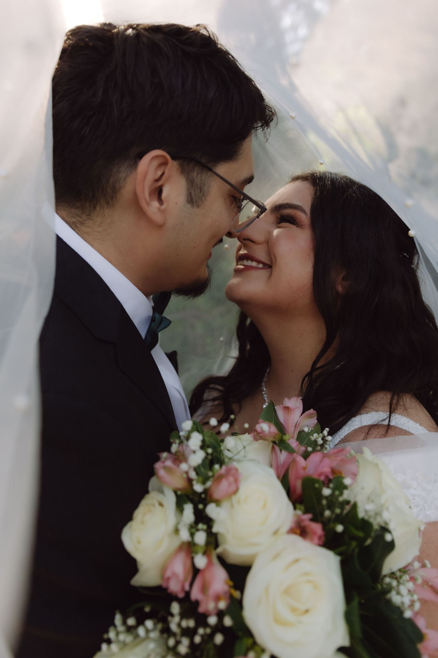 Smiling bride and groom touch noses under a wedding veil, bride holding a white and pink bouquet.