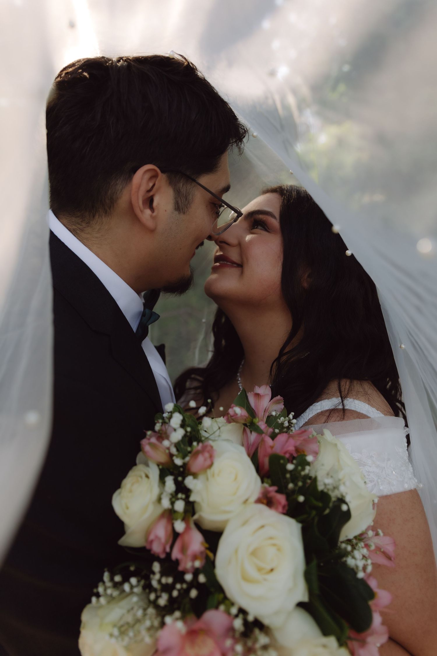 Bride smiles joyfully at groom under a flowing wedding veil while holding a bouquet of white roses.