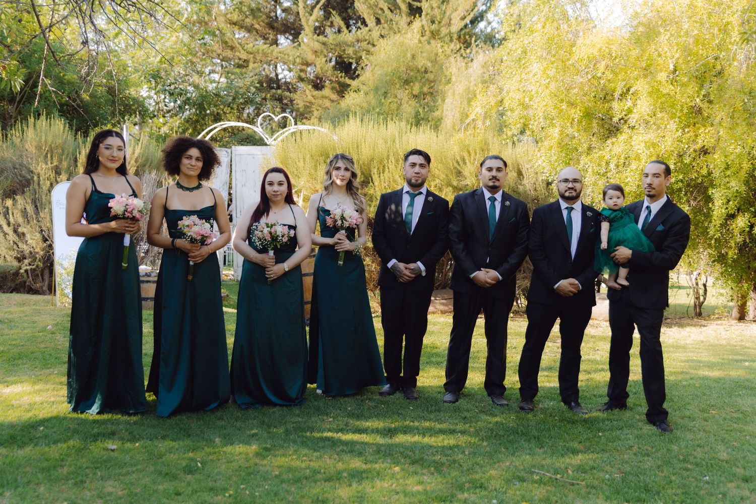Wedding party poses outdoors with bridesmaids in emerald green gowns and groomsmen in black suits.