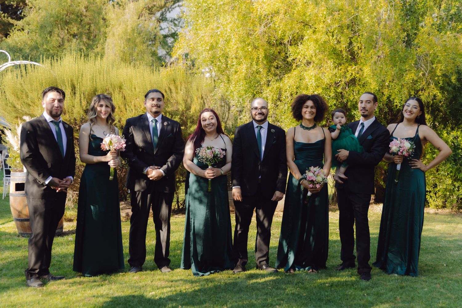 Wedding party group photo on green lawn with bridesmaids in dark green dresses and groomsmen in suits.