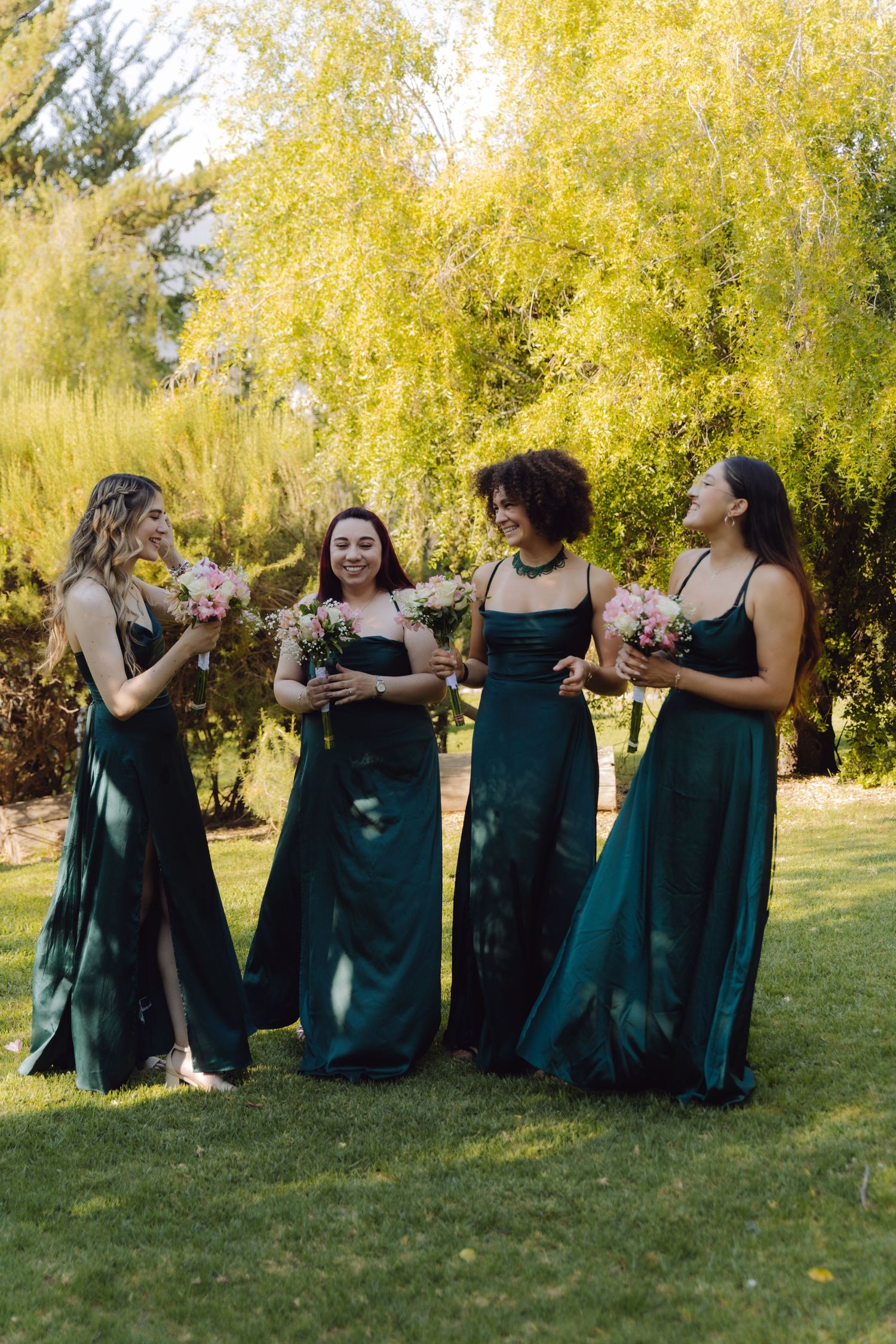 Four bridesmaids in emerald green gowns laughing and holding pink bouquets outdoors at a wedding.