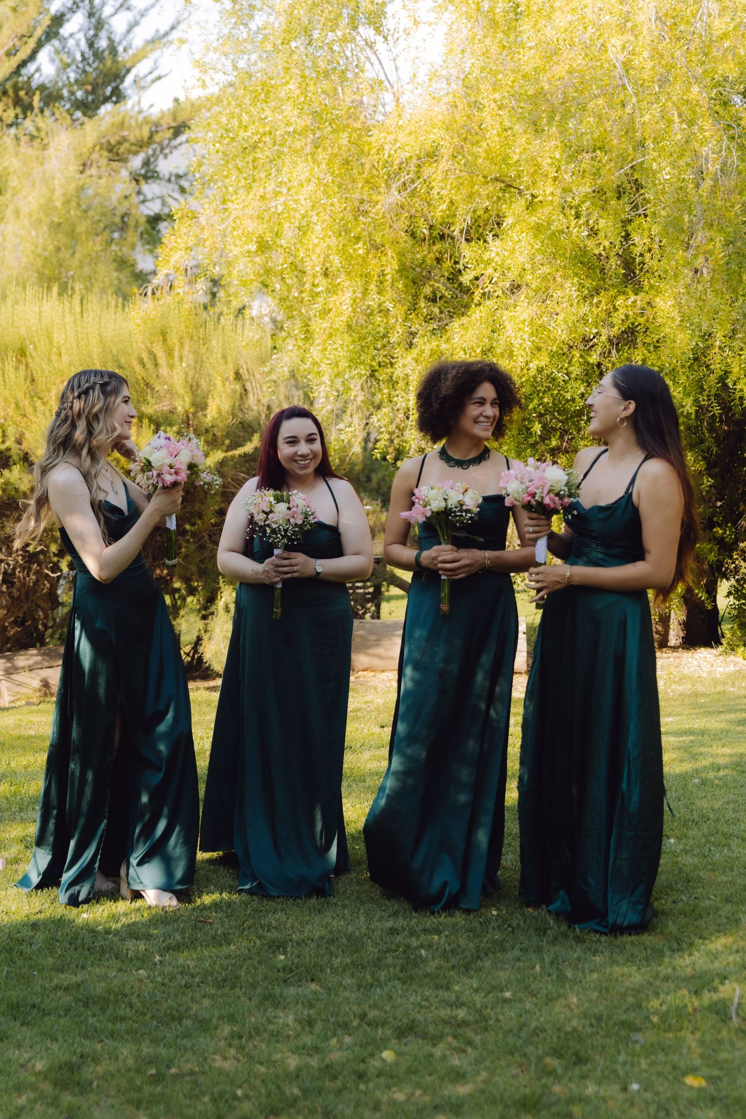 Four bridesmaids wearing matching dark green satin dresses holding pink floral bouquets at outdoor wedding.