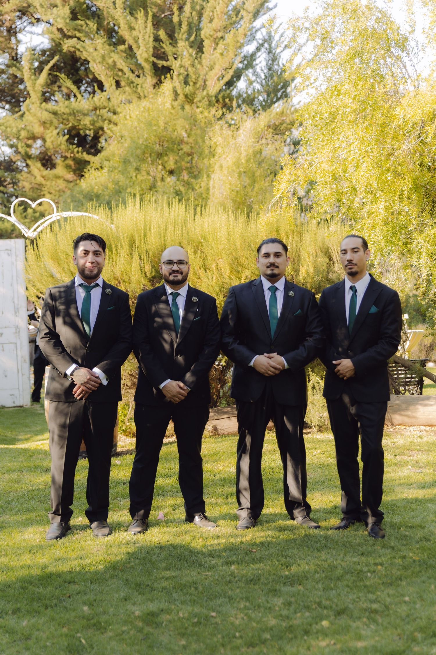 Four groomsmen in formal dark suits with green ties standing together at outdoor garden wedding.