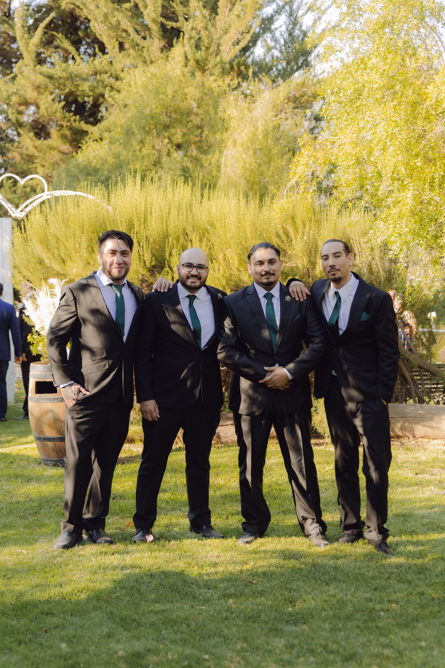 Four groomsmen in dark suits with green ties posing together on grass at outdoor wedding venue.