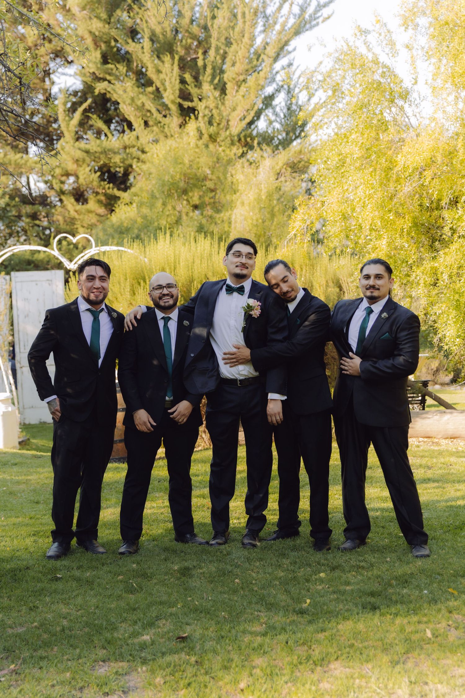 Five groomsmen in black suits laughing and posing playfully together at outdoor garden wedding ceremony.