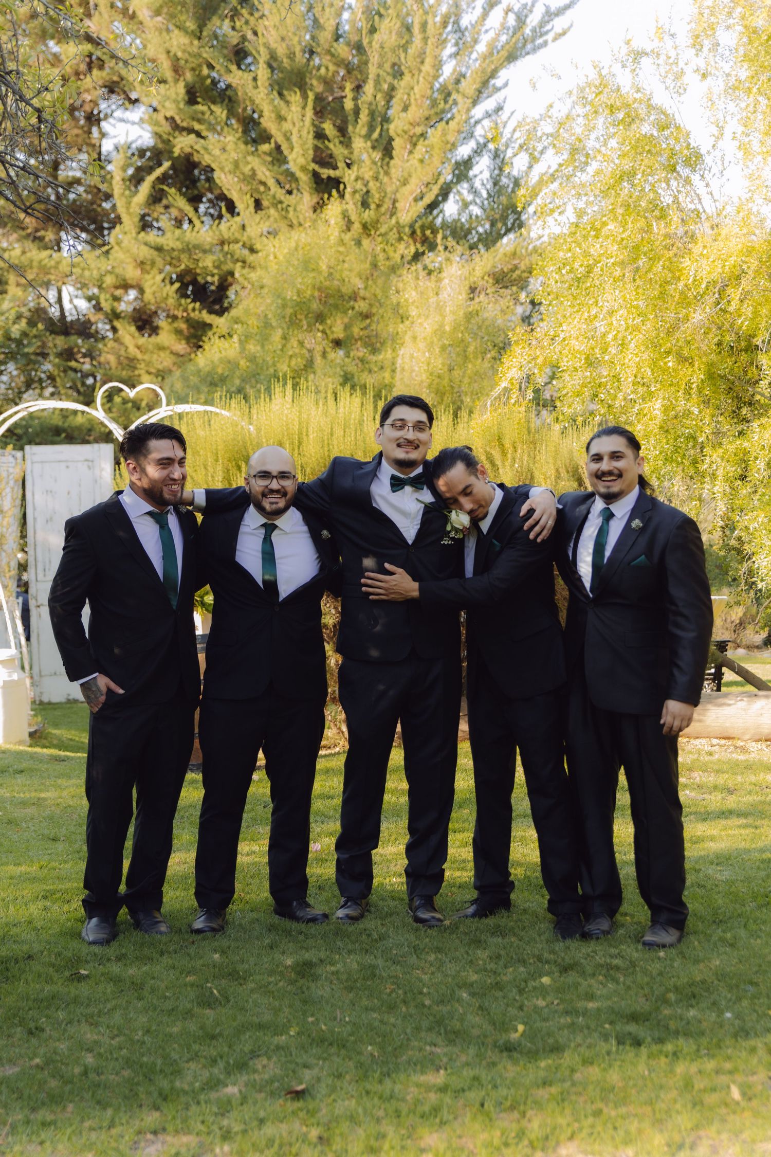 Five groomsmen in black suits with green ties smiling together at outdoor garden wedding venue.