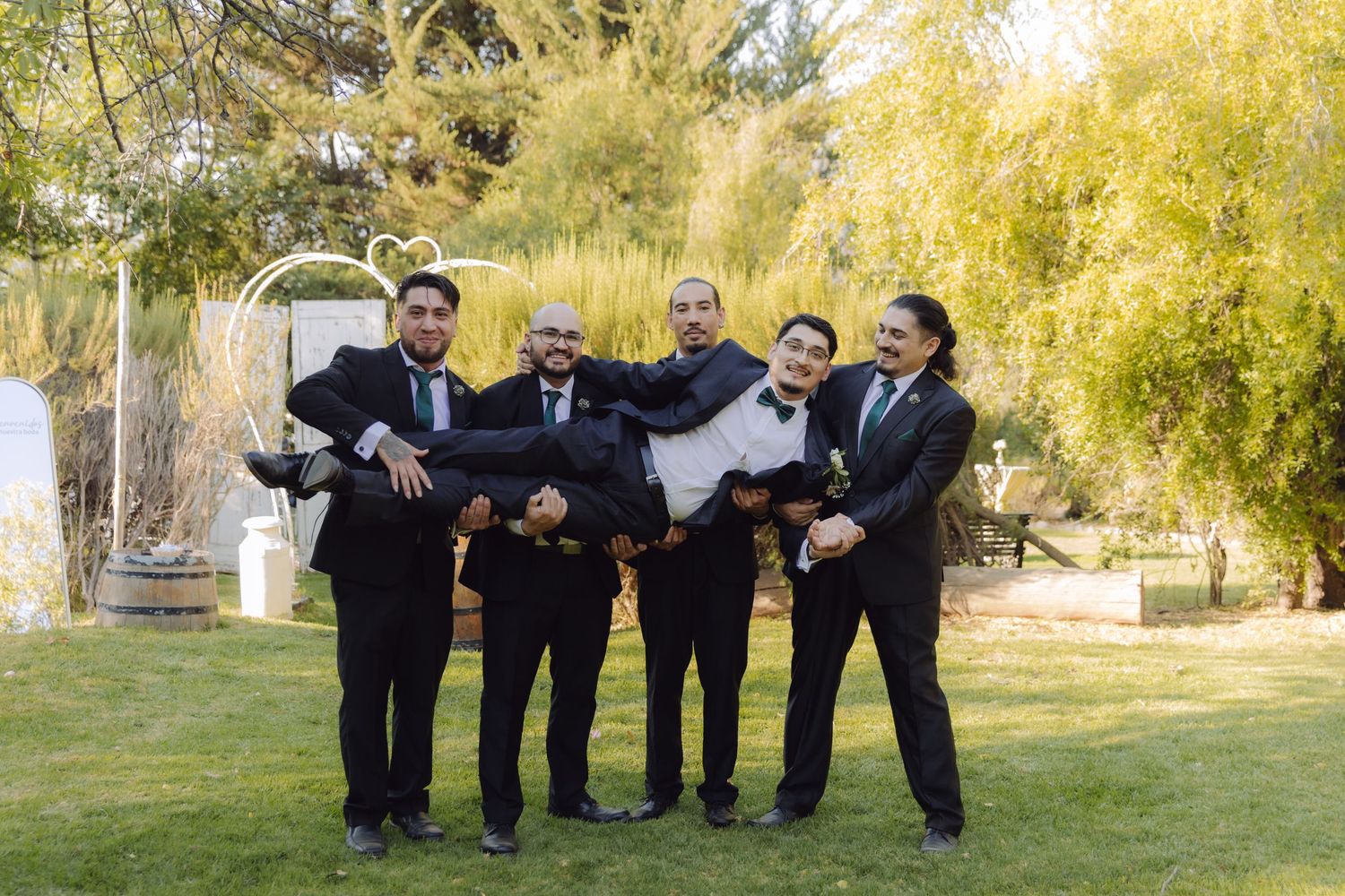 Four groomsmen lifting groom in black tuxedo playfully at outdoor garden wedding celebration.