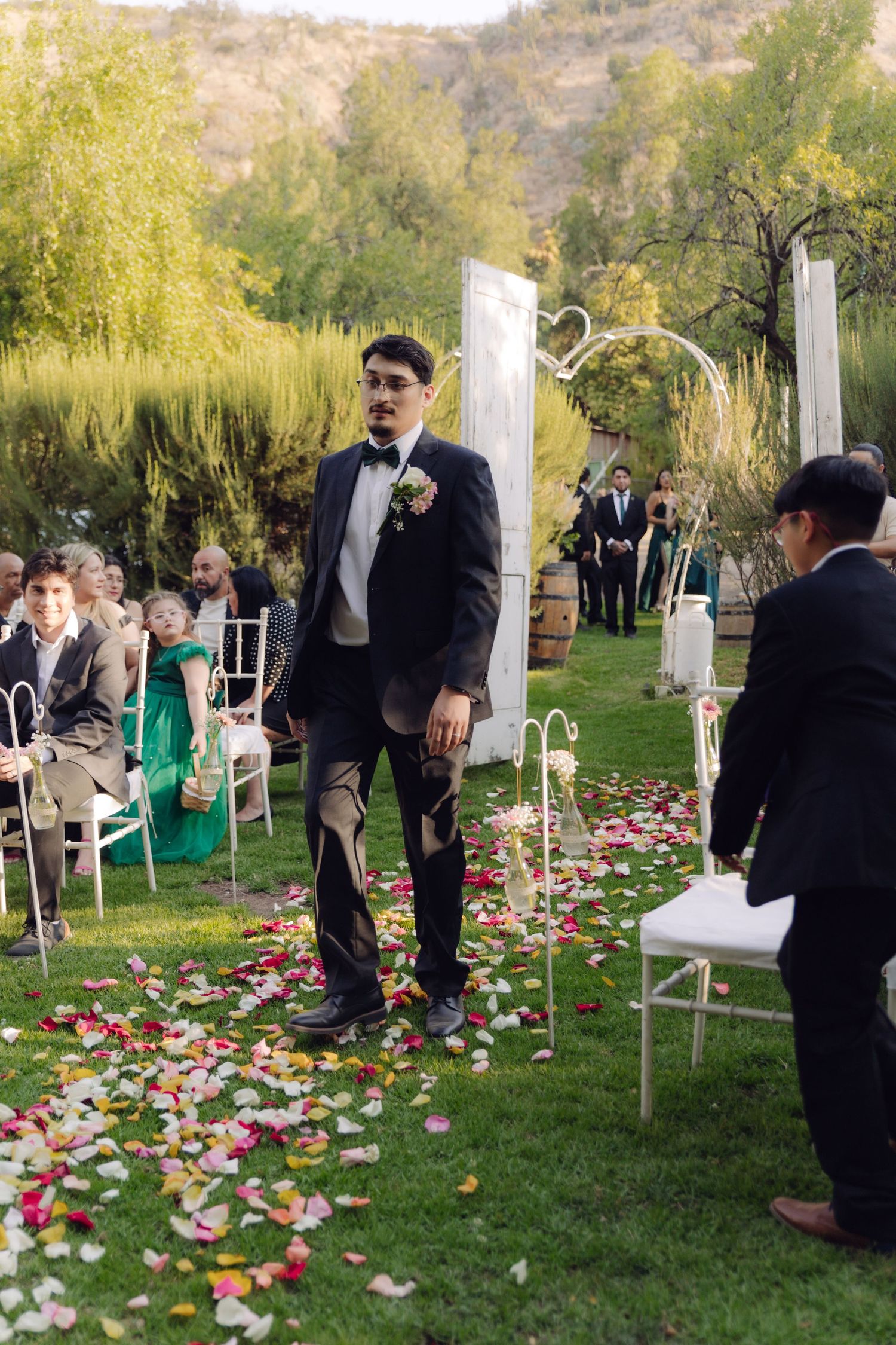 Groom in black tuxedo walking down petal-lined aisle at elegant outdoor garden wedding ceremony.