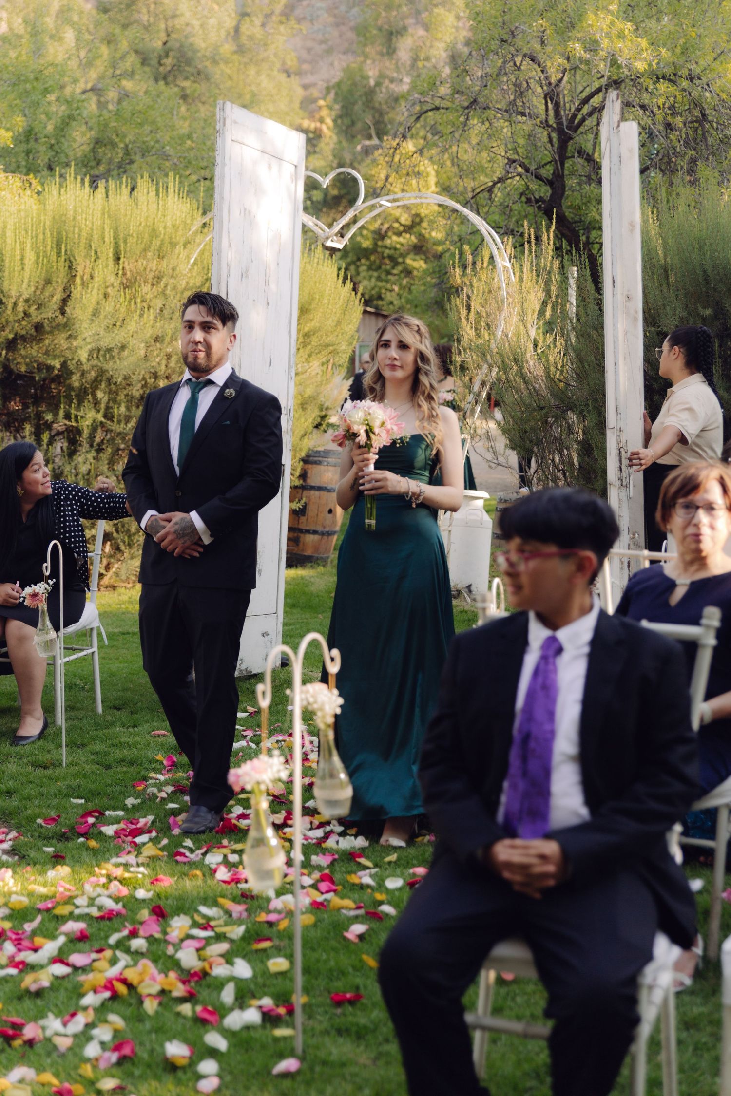 Groomsman escorts bridesmaid in green gown down rose petal aisle at outdoor wedding ceremony.