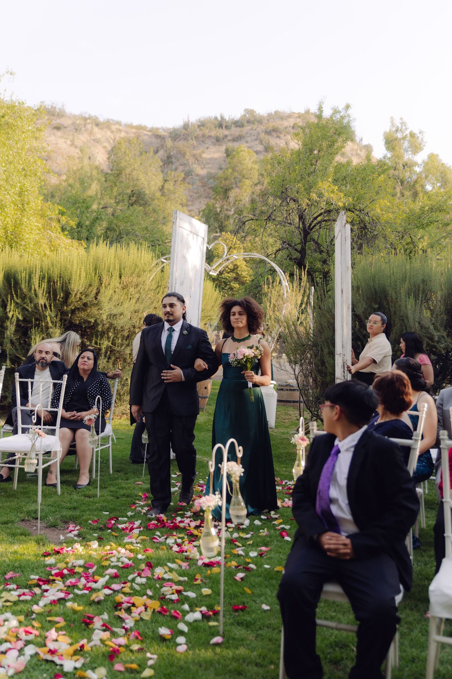 Groomsman and bridesmaid in emerald dress walking down petal-lined aisle at outdoor mountain wedding.