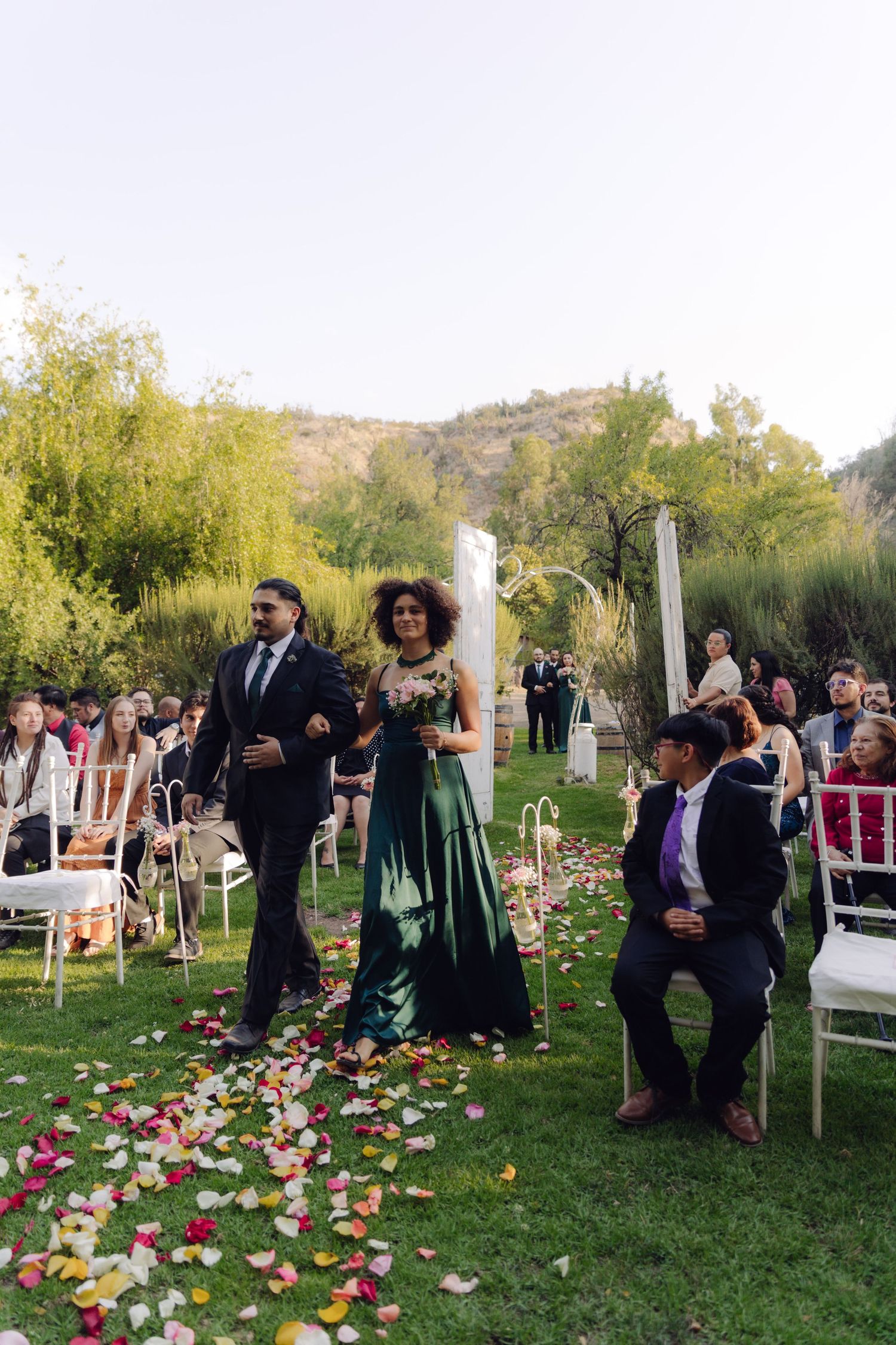Bridesmaid in emerald green gown walks down petal-strewn aisle at outdoor wedding ceremony.