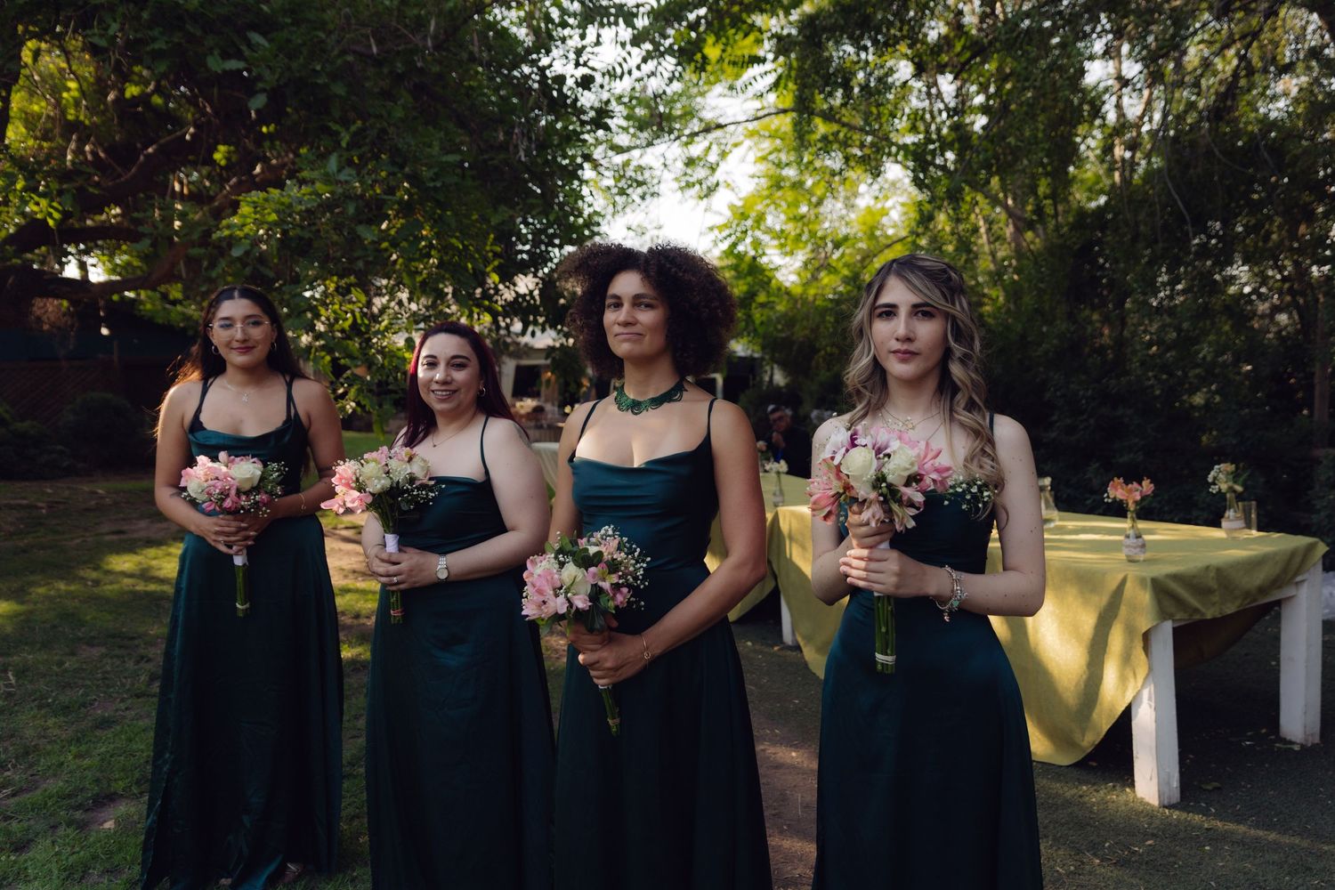 Four bridesmaids wearing elegant dark green dresses holding floral bouquets outdoors.