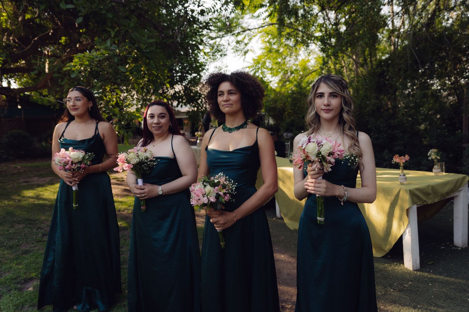 Four bridesmaids in matching dark green gowns holding bouquets at outdoor wedding.