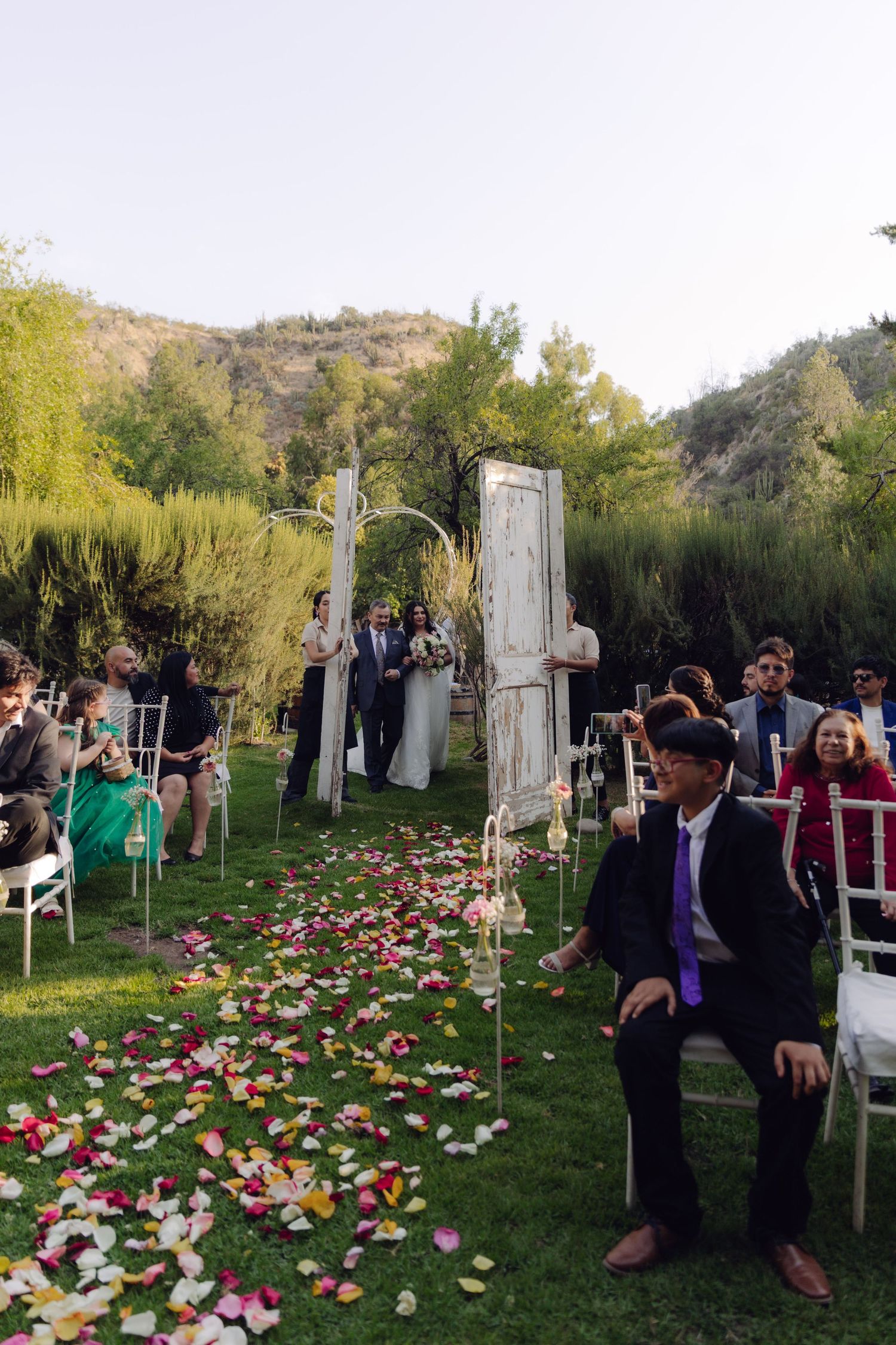 Scenic outdoor wedding ceremony with petal-lined aisle, white doors, and mountain backdrop.