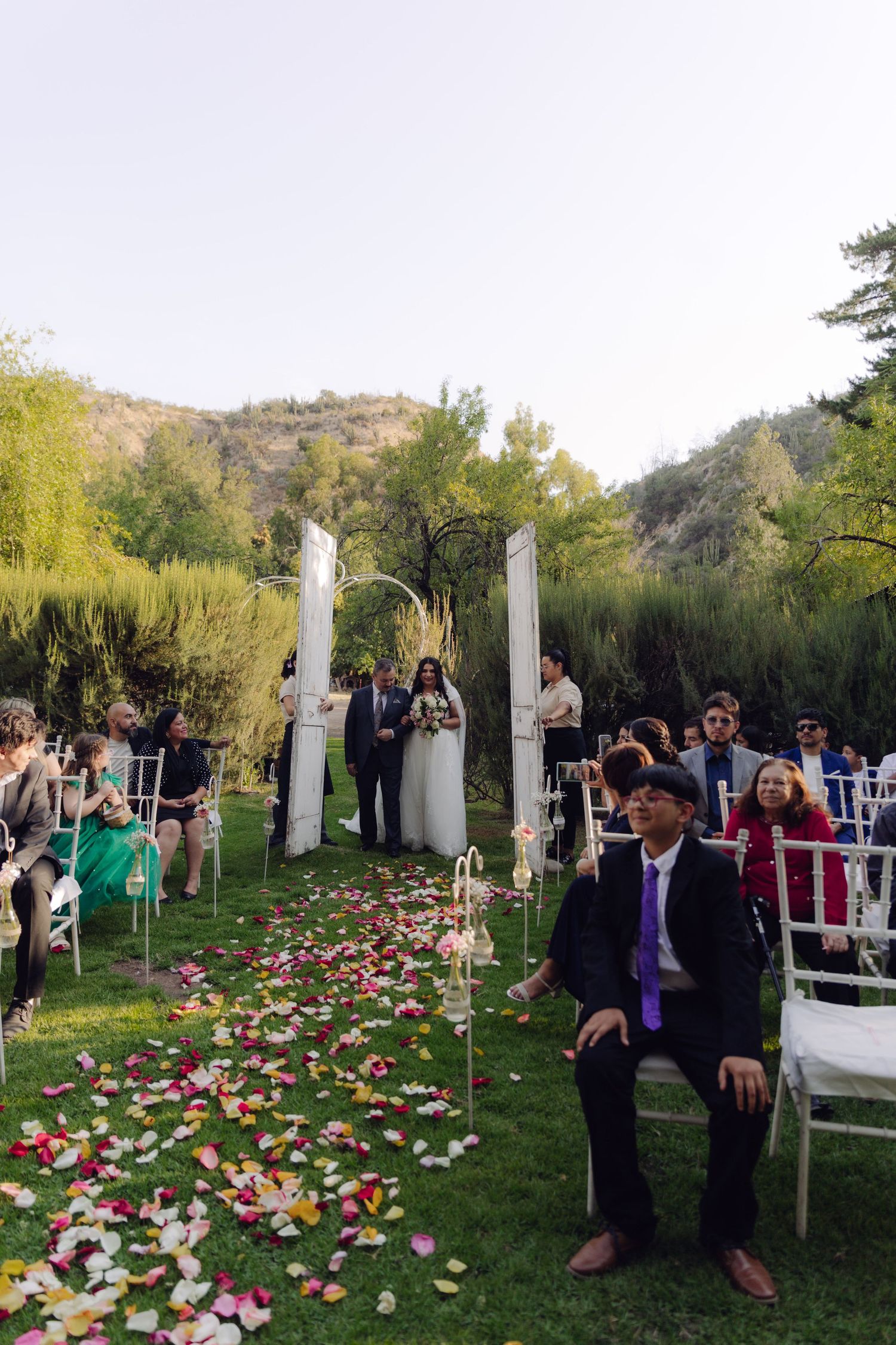 Outdoor wedding ceremony aisle with colorful rose petals and white chairs on green lawn.