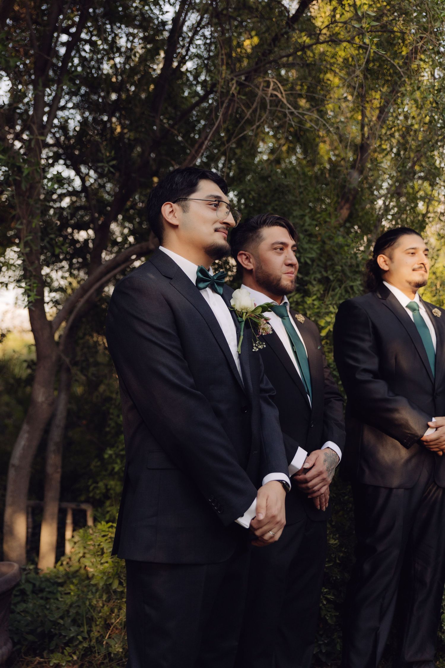 Three groomsmen in black tuxedos with green bow ties standing outdoors at wedding.