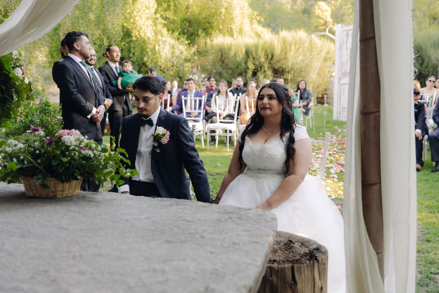 Bride and groom seated together at outdoor wedding ceremony under floral arch.