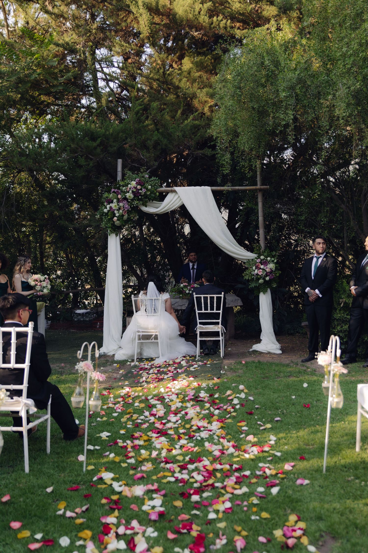 Elegant outdoor wedding altar with white draping, floral arch, and rose petal aisle.
