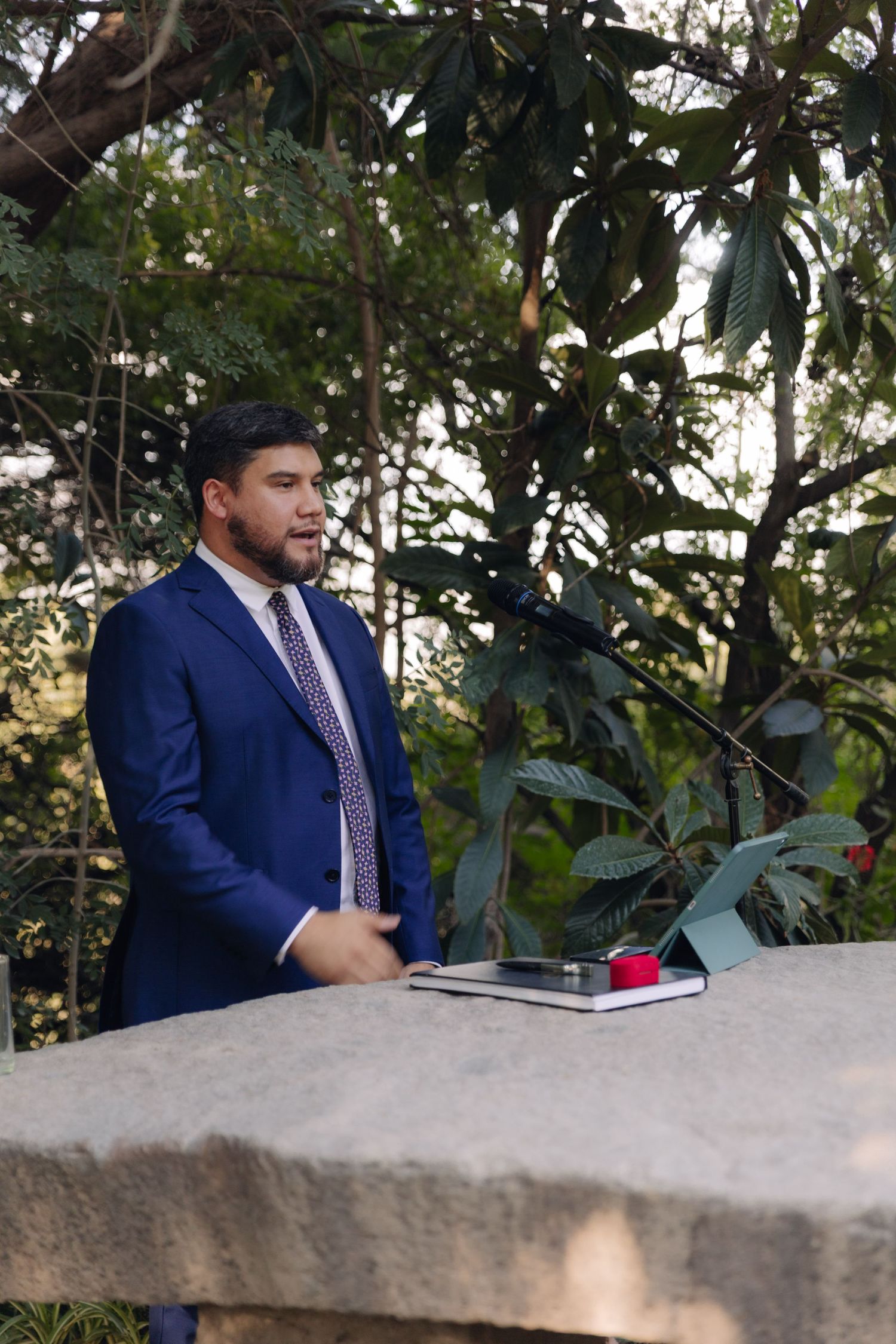 Man in blue suit speaking at outdoor podium with microphone surrounded by lush green trees.