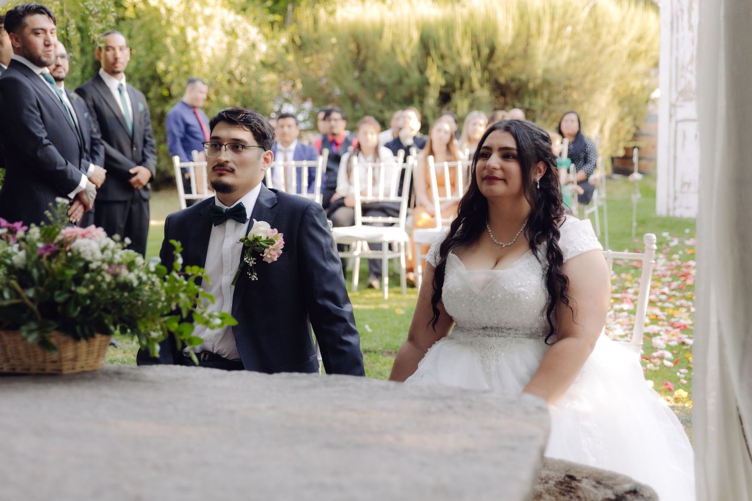 Bride and groom kneel together during outdoor wedding ceremony under floral canopy.