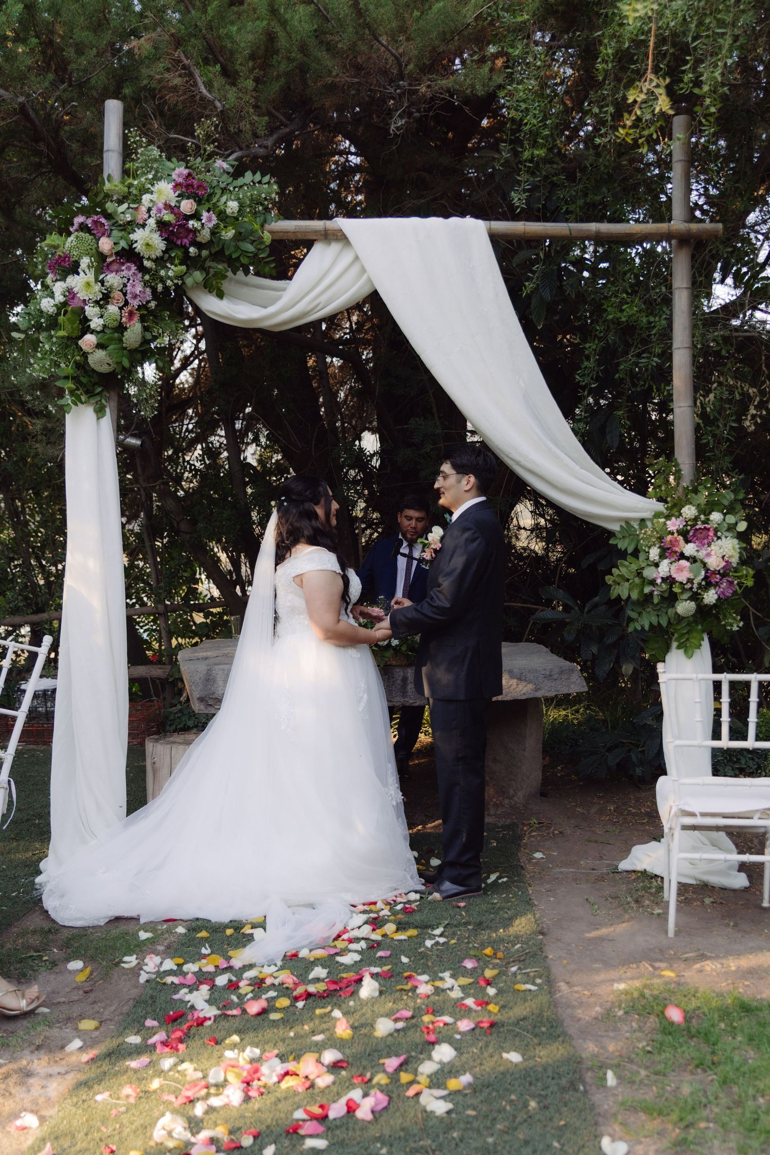 Bride and groom exchanging vows under floral arch with white draping and rose petals.