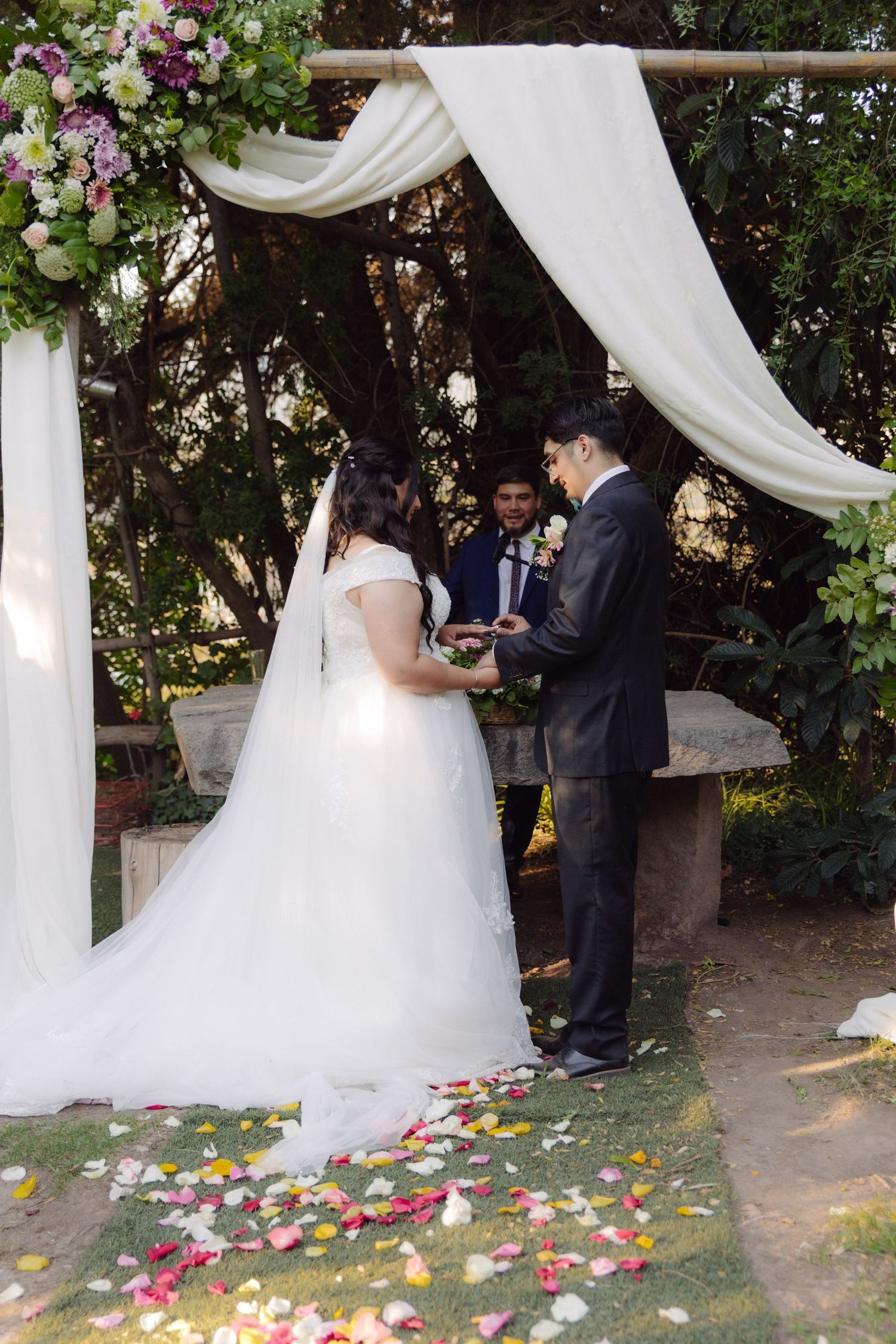 Couple holding hands at outdoor wedding altar decorated with flowers and white fabric draping.
