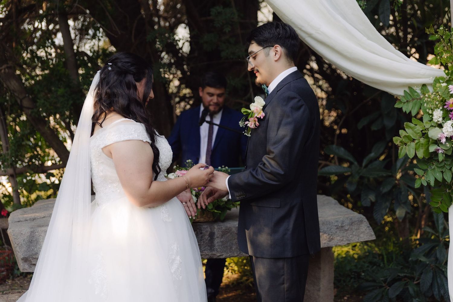 Bride and groom exchanging rings under floral wedding arch in garden ceremony setting.