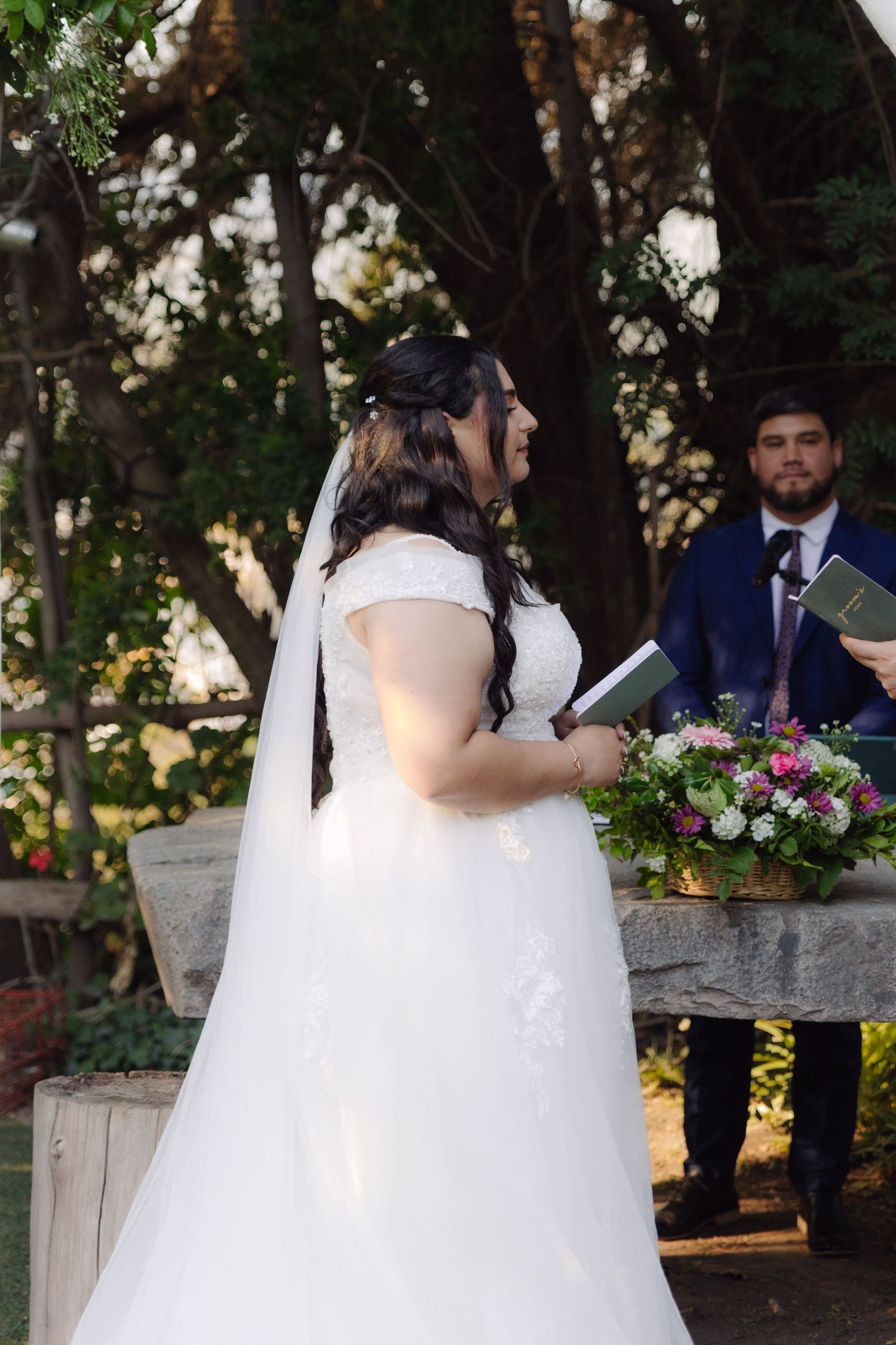 Bride in white gown smiling during outdoor garden wedding ceremony with officiant nearby.